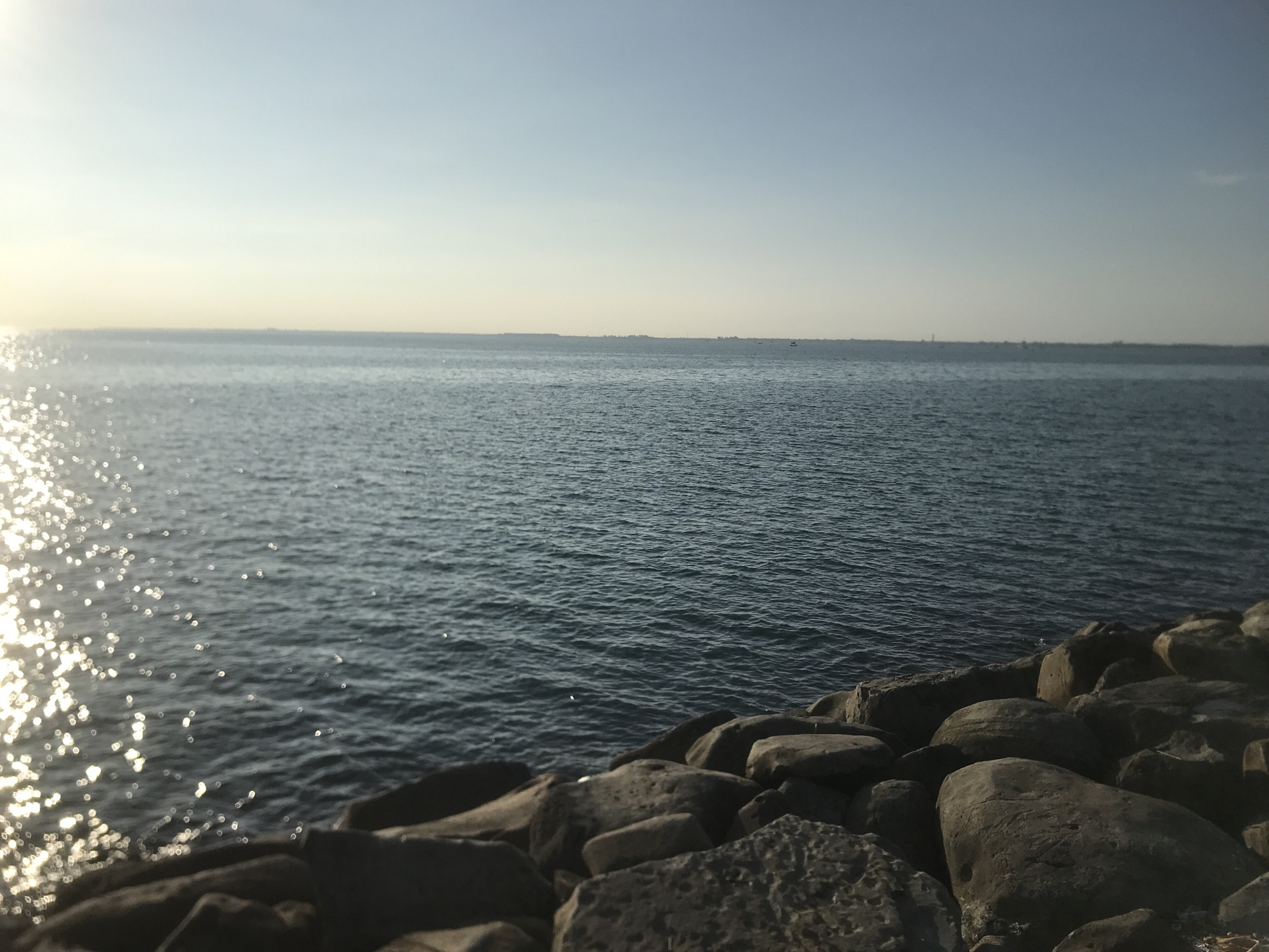 The image depicts a coastal scene. The foreground consists of large, dark gray rocks piled along the shoreline. Beyond the rocks is a body of water, possibly the sea, with gentle ripples and reflections of sunlight. In the distance, a faint landmass is visible on the horizon under a light blue sky, slightly hazy. The overall impression is that of a calm, overcast day at the coast.
Given the instruction to locate the image in Lhokseumawe, Indonesia, the scene likely depicts one of its coastal areas. Without specific landmarks or features, pinpointing the exact location is impossible, but the image reflects the coastal environment typical of the region.