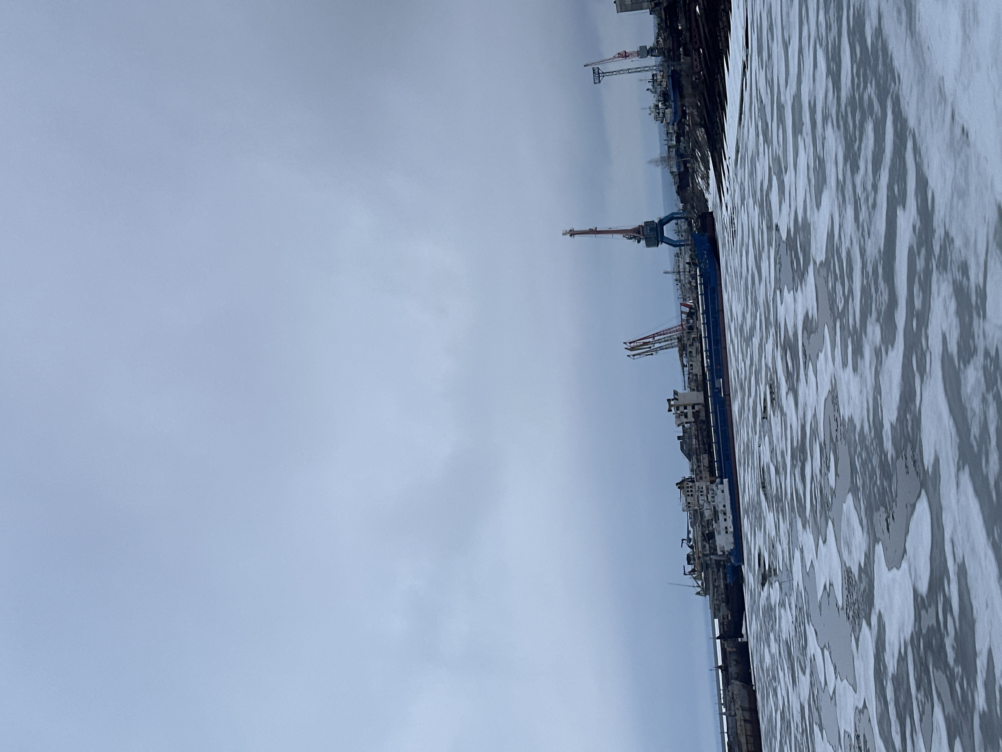 The image depicts a harbor scene in Chaikovsky, Russia. The sky is overcast. Several gantry cranes are visible on the waterfront. Cargo ships are docked alongside the pier. Cracked ice floats on the water's surface in the foreground.