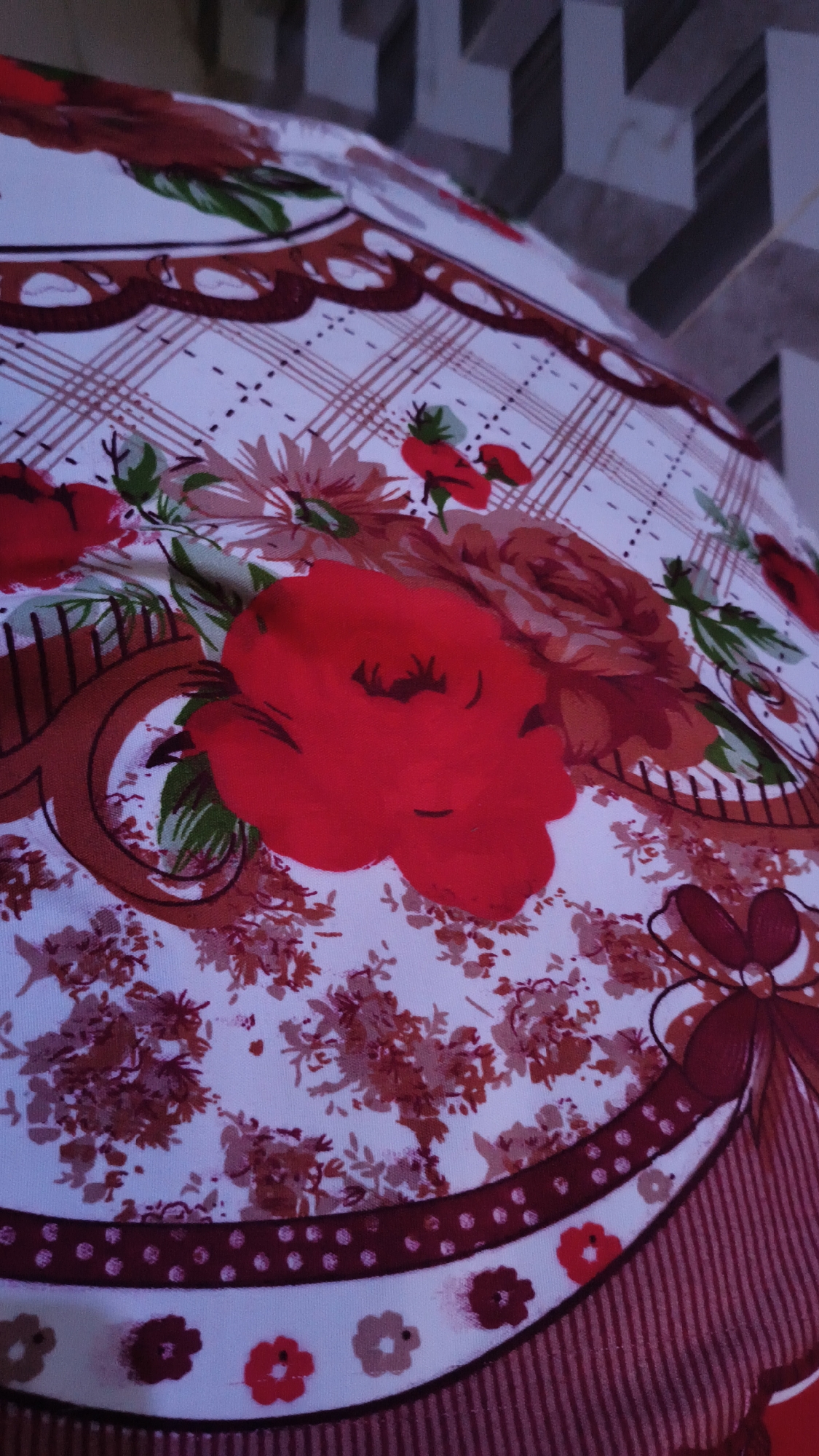 The image shows a close-up of a round, floral patterned tablecloth or fabric. The pattern features large red and pink flowers, green leaves, and a brown and white checkered design with red lines. There are also decorative brown borders with scalloped edges and a red bow. The fabric appears to be laid out on a surface, with part of a beige and grey tiled wall visible in the background, suggesting an indoor setting. The lighting is somewhat dim. There are no people or actions depicted in the image.