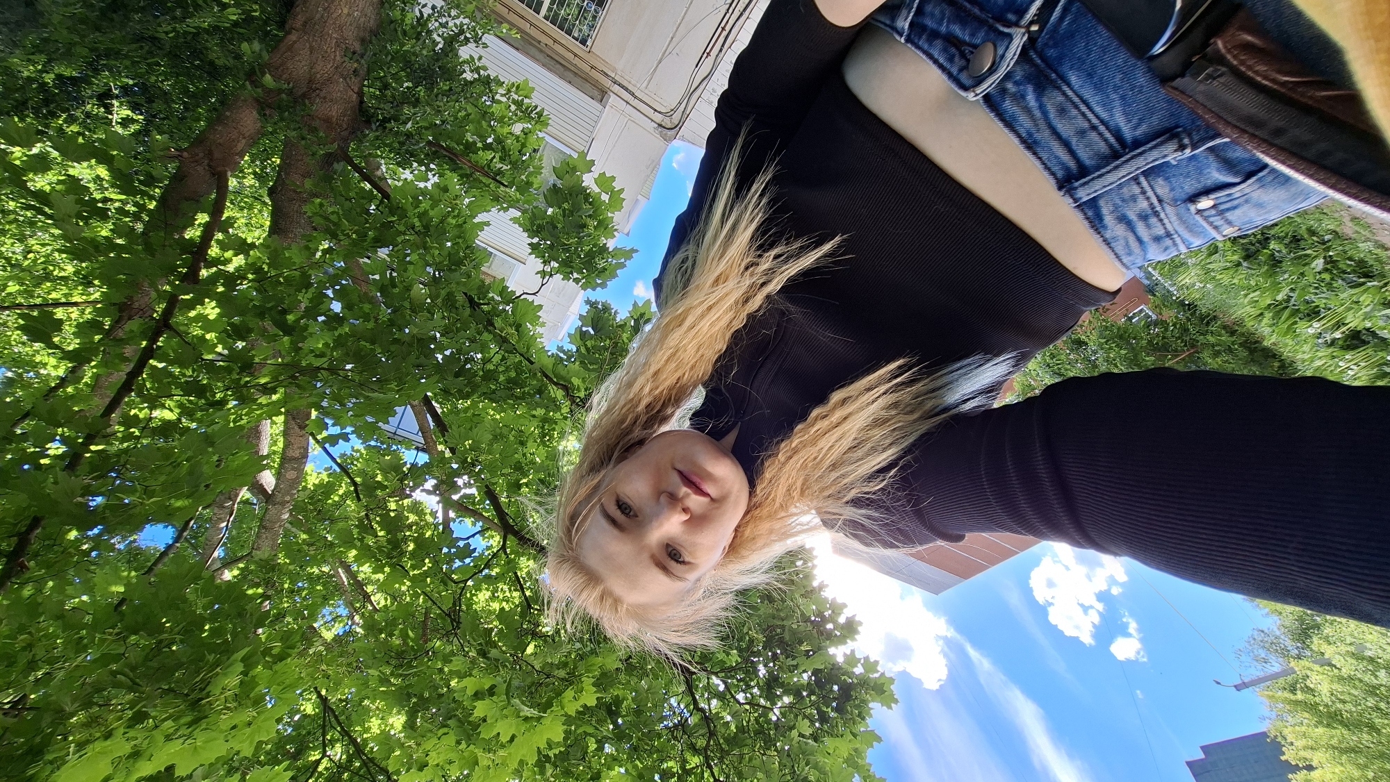 A woman with long blonde hair, wearing a black long-sleeved shirt and blue denim shorts, is positioned in the center of the frame. The image is taken from a low angle, looking up at her. Above and around her are green tree leaves and branches against a background of blue sky with white clouds. A building can be seen in the upper left of the frame. The photo seems to have been taken in an outdoor setting, potentially a park or garden, within the city of Chaikovsky, Russia.