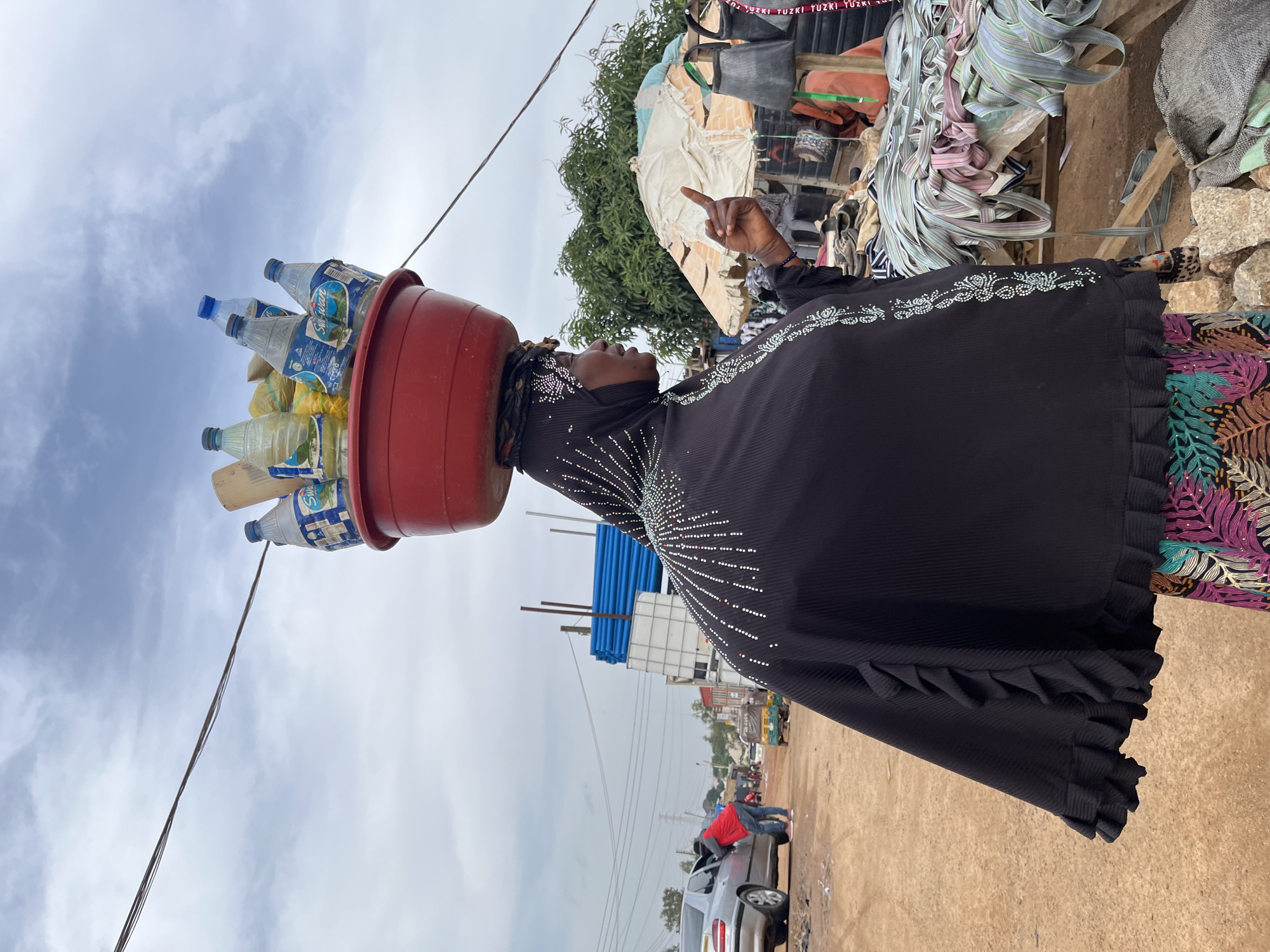 A woman in a black dress adorned with white beading stands in an outdoor market, balancing a red basin filled with plastic water bottles on her head. Her right hand is raised, pointing upwards with her index finger. The scene appears to be a busy marketplace, with various goods and structures visible in the background, including what looks like a stall with textiles and other items. The sky is cloudy, suggesting it could be daytime with overcast weather. The ground is dusty, characteristic of a dry, outdoor environment. The presence of a silver car and other vehicles in the background indicates a public area, likely a road or a part of the market accessible by vehicles. The woman's gesture and the way she carries the water bottles suggest she is engaged in selling or transporting them, possibly at a market in Gura Top, Nigeria. The overall impression is one of daily life and commerce in a bustling community.
