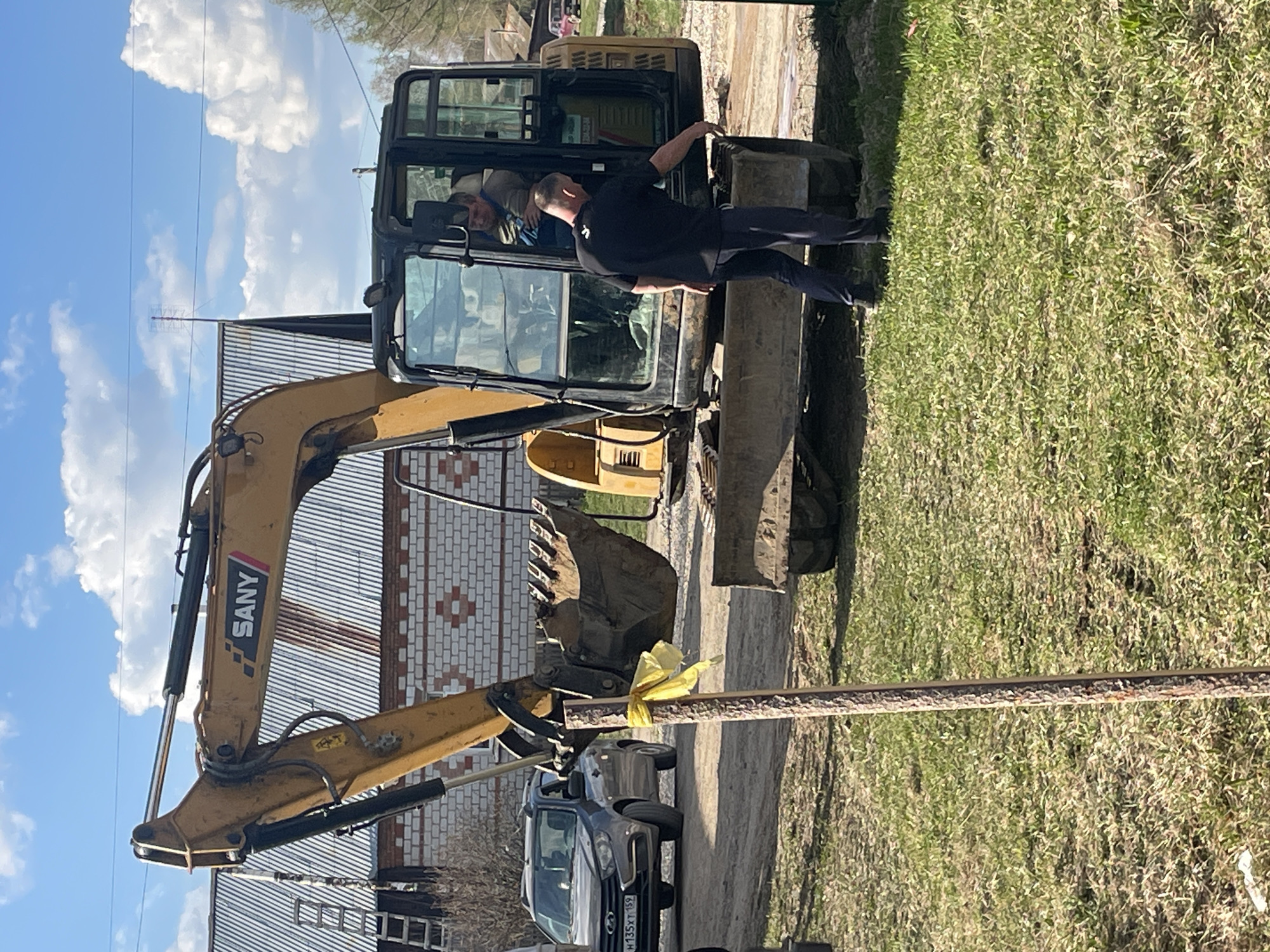 The scene is set outdoors in Поедуги, Russia. An excavator with the logo "SANY" visible on its arm is prominently displayed. A log is tied to the excavator's arm with a yellow ribbon. A person wearing a black shirt and dark pants is standing alongside the excavator's cabin, with another person visible inside the cabin. Behind the excavator, there is a building with a corrugated metal roof and brickwork on the lower part. In the foreground, a grey car is parked, its license plate partially visible as "H135XT." Grass and sky with clouds are also visible in the background.