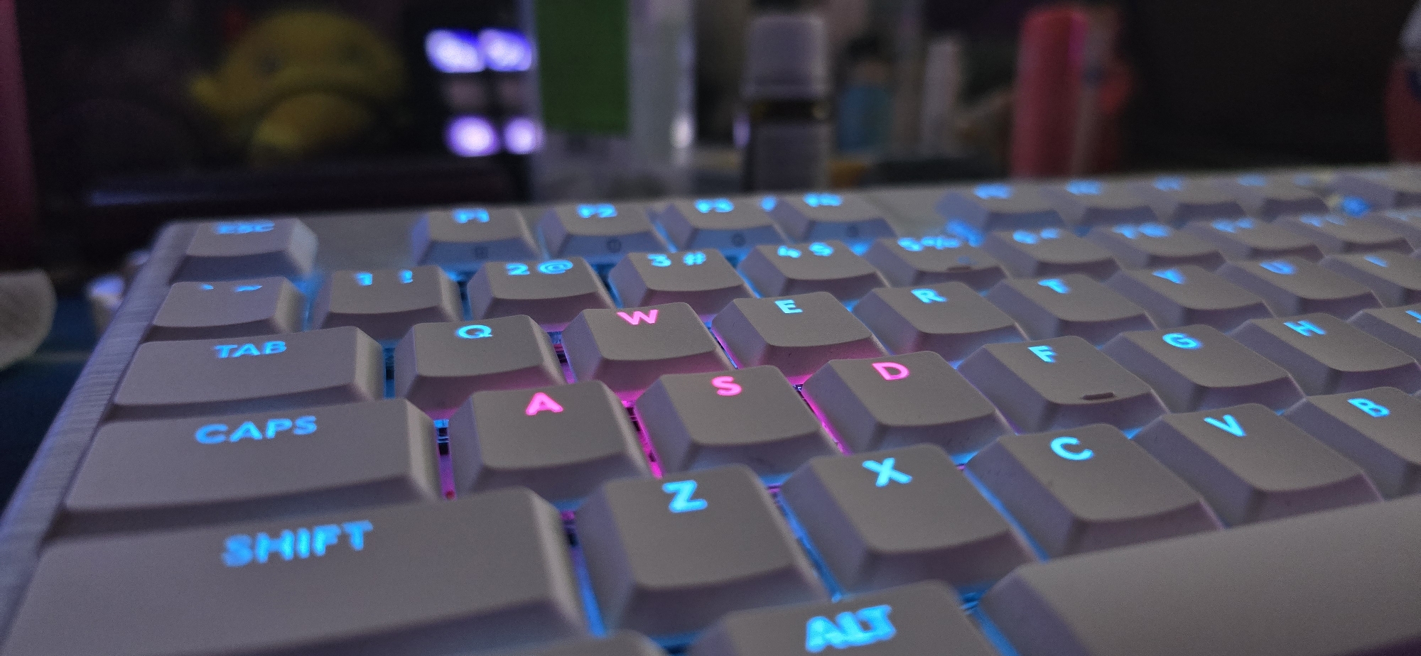 A close-up, low-angle shot captures a white, backlit mechanical keyboard with a shallow depth of field, blurring the background. The keyboard features keycaps illuminated in cyan/blue and magenta/pink, creating a vibrant RGB effect.
Visible keys include "TAB," "CAPS," "SHIFT" (all in cyan/blue), and parts of the QWERTY layout. Keys like 'Q', 'W', 'E', 'R', 'A', 'S', 'D', 'F', 'G', 'H', 'Z', 'X', 'C', 'V', 'B', and "ALT" are discernible. The 'A', 'S', 'D', and 'W' keys are distinctly illuminated in magenta/pink, suggesting they might be configured as common gaming keys, while others are cyan/blue. Numbers '1', '2@', and '3#' are also visible on the top row, glowing in cyan/blue.
The background is heavily blurred but hints at indoor surroundings, possibly a desk setup. A yellow, duck-like plush toy is vaguely visible in the top-left background, along with indistinct bottles or containers and blurred light sources, suggesting dim ambient lighting, likely evening or night. The overall scene conveys a relaxed, tech-oriented environment in Burgas, Bulgaria, ready for typing or gaming.
