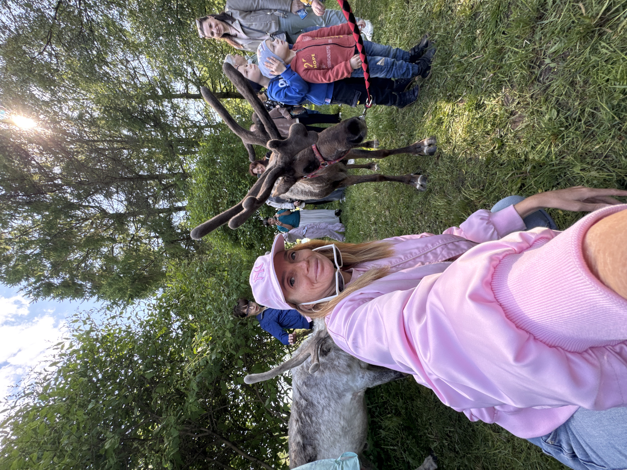 A woman in a pink jacket and baseball cap is taking a selfie with a reindeer in Vsevolozhsk, Russia. The reindeer has large antlers and is wearing a red collar. Behind the reindeer, a group of people, including children, are standing in line. The background is a green grassy area with trees. The weather is sunny.