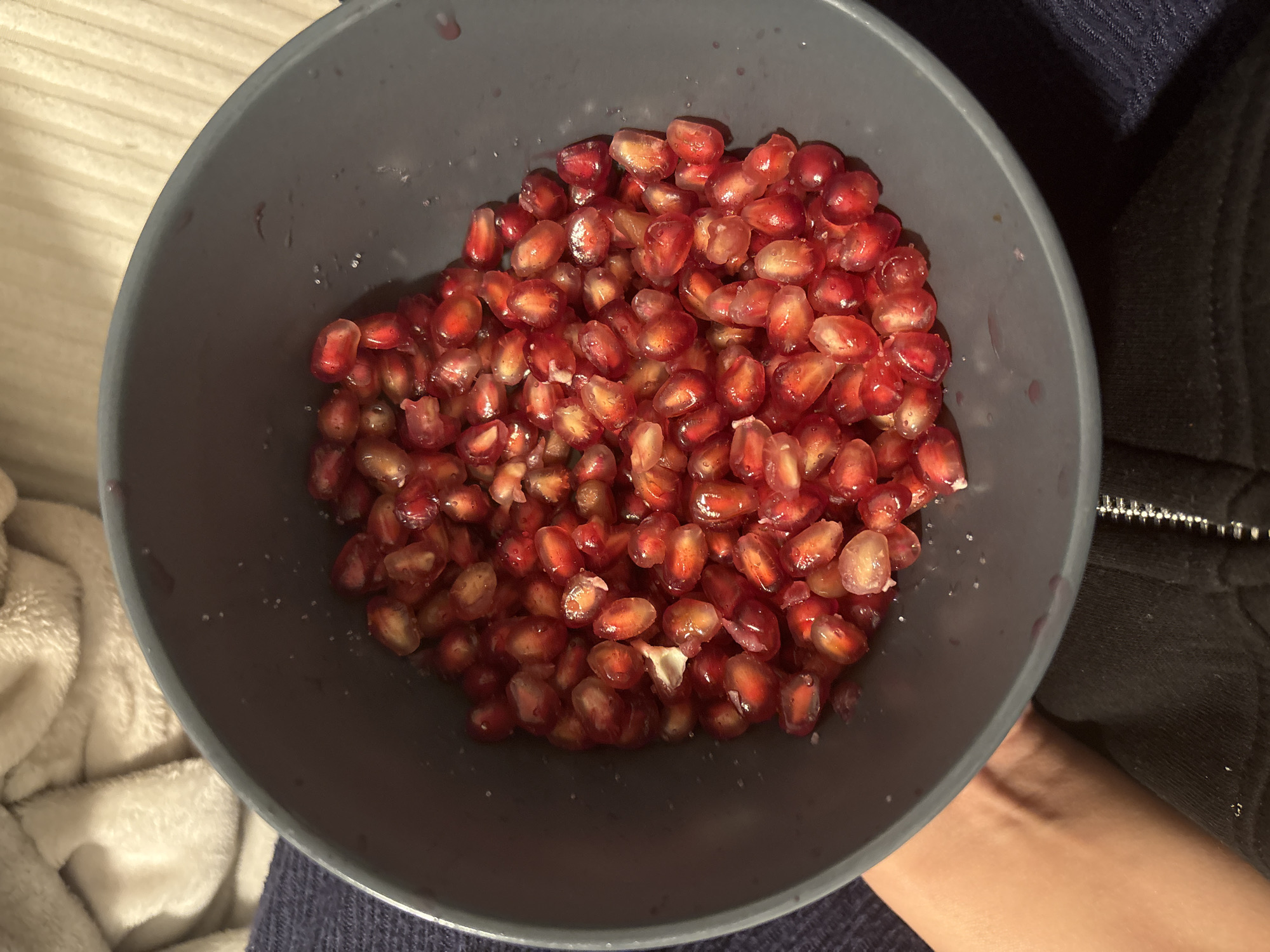 In a grey bowl, a generous portion of pomegranate seeds is displayed. The seeds are plump and juicy, with a vibrant red hue and some translucent qualities. They are piled high, some seeds overlapping others, creating a textured surface within the bowl. A few small water droplets and specks of what appears to be sugar are visible amongst the seeds. The bowl itself has a matte finish and a slightly textured interior. The background is softly blurred, showing hints of a light-colored, fuzzy fabric and a dark blue textured material. A person's hand and arm are partially visible on the left side of the image, appearing to hold the bowl. The scene is indoors, with the lighting suggesting an ambient light source, possibly from above or the side. There are no discernible landmarks or elements that place this image specifically in Mönchengladbach, Germany; it is a generic domestic scene.