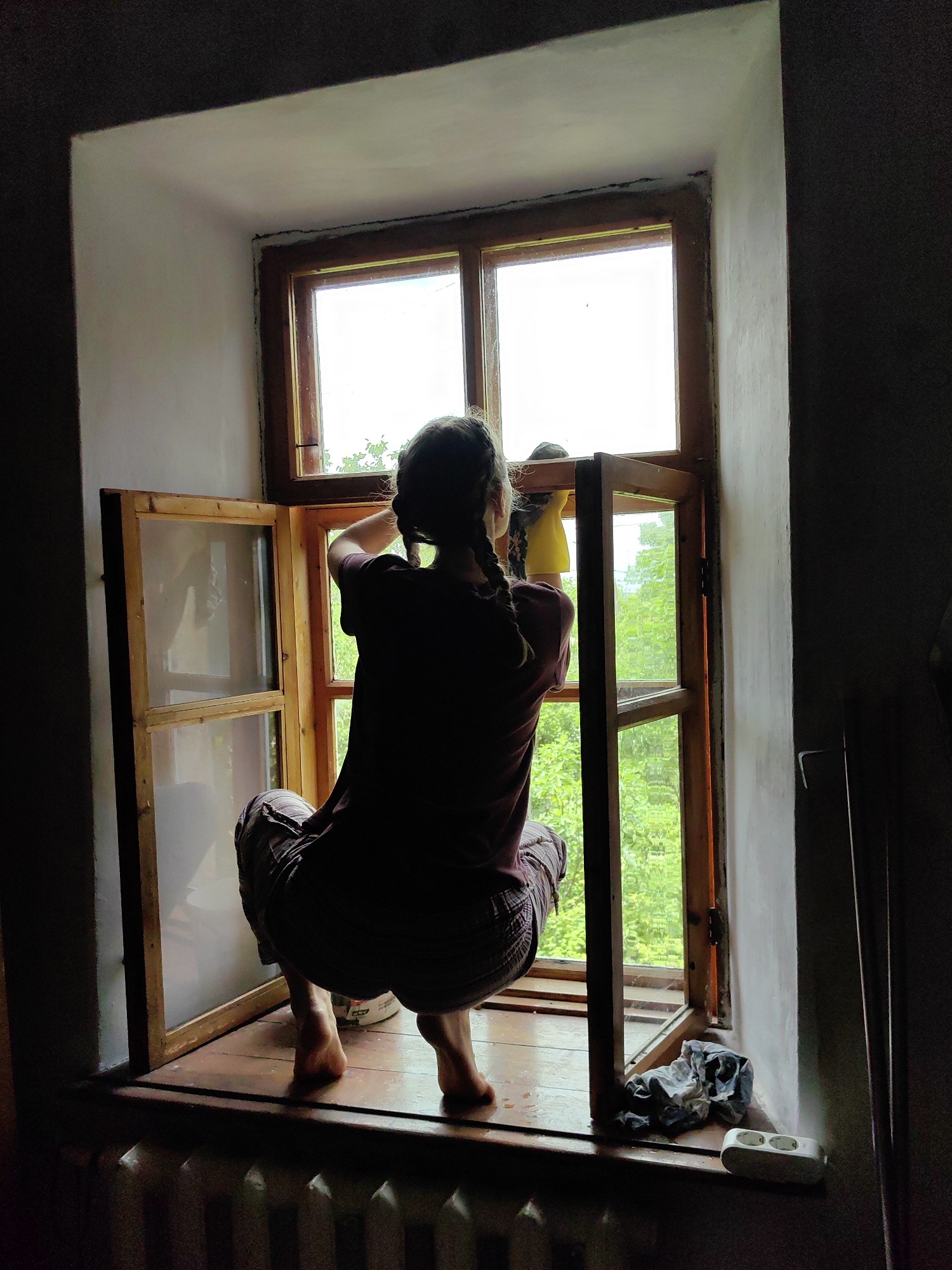 An adult individual, seen from behind, is squatting barefoot on a wooden windowsill, actively cleaning a window. The person has dark hair styled in two braids and is wearing a dark red short-sleeved top and dark, striped trousers. Their right hand, covered by a yellow glove, holds a dark cloth and is wiping the upper right pane of a multi-pane wooden window.
The window features multiple wooden frames, with one section on the left open inwards and another section on the right seemingly open outwards or removed for access. Bright daylight illuminates the scene from outside, revealing green foliage through the lower window panes and a bright white sky above.
The interior wall surrounding the window is off-white and shows signs of wear, with dark lines visible along the juncture with the wooden window frame. The wide wooden windowsill is wet in places. To the person's left, a white bucket or container is on the sill. To the right, a crumpled multi-colored cloth and a white electrical power strip with two outlets are positioned on the sill. A white, ribbed radiator is partially visible beneath the windowsill. The room's interior is dimly lit, silhouetting the person against the bright outdoor view. The scene takes place in a residential setting in the city of Unknown, Unknown.