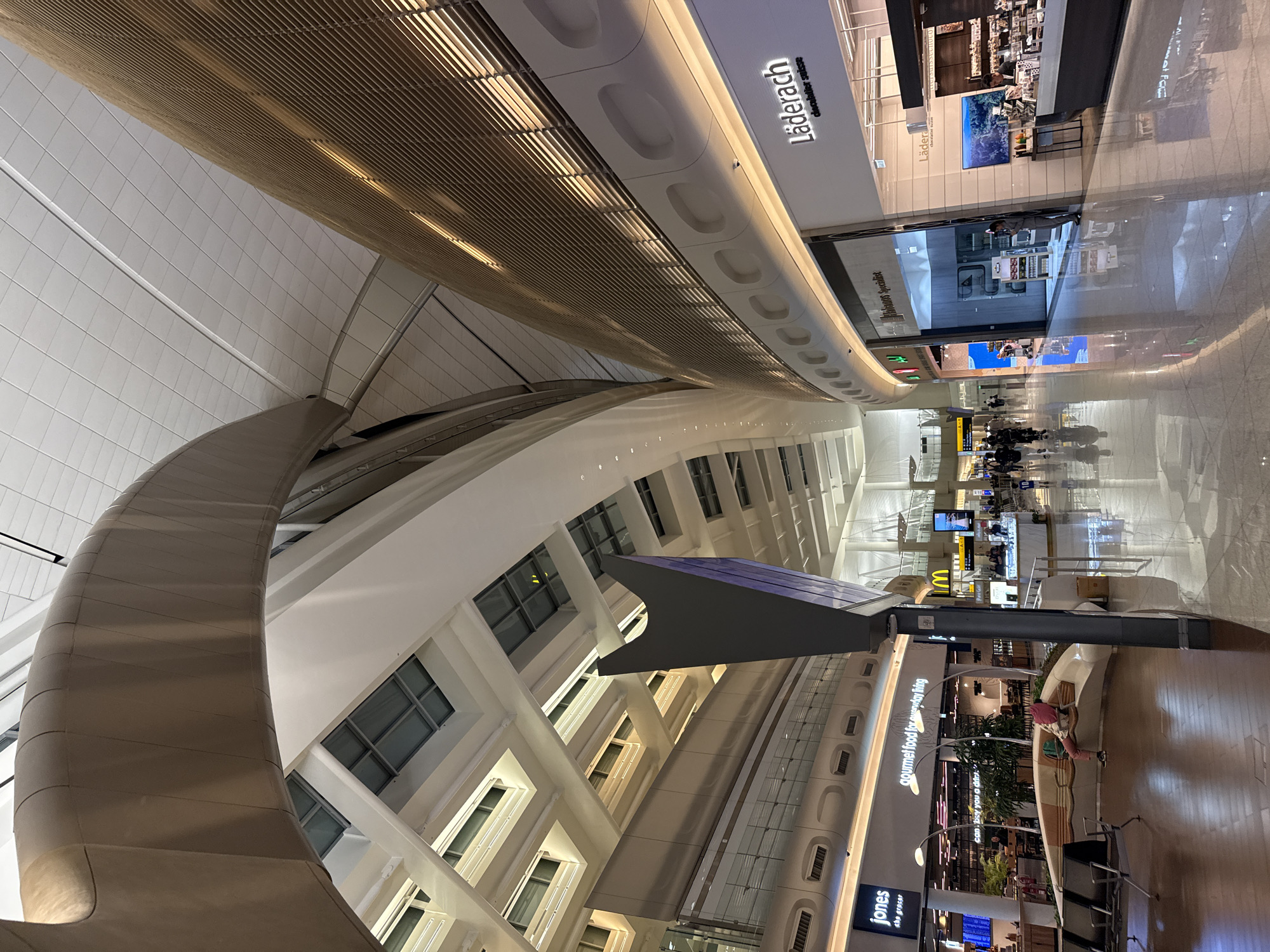 The image shows the interior of a modern airport terminal. The architecture is characterized by large, curved structures and a bright, airy atmosphere, with a tiled ceiling and large windows or skylights. In the foreground, there is a polished floor reflecting the ambient light.
Several shops and businesses are visible along the concourse. To the left, there is a shop labeled "jones the grocer" and another with the text "gourmet food for everyday living." Further down the concourse, there is a McDonald's restaurant. To the right, there are storefronts, including one clearly labeled "Läderach chocolatier suisse," which appears to be a chocolate shop.
There are a few people visible in the terminal. In the left foreground, a person is sitting on a curved bench, wearing a pink head covering and pink pants, with a backpack next to them. Other people are walking or standing in the background, suggesting a busy public space.
The overall environment is clean and well-lit, typical of an international airport terminal. The lighting is a mix of overhead artificial lights and natural light coming from the upper sections of the terminal. The scene appears to be in a public area of the airport, possibly a departure or arrival hall. Without further information, it's not possible to definitively place this in Al Matar, United Arab Emirates, but the modern design and international branding of the shops suggest it could be a major airport in a developed city.