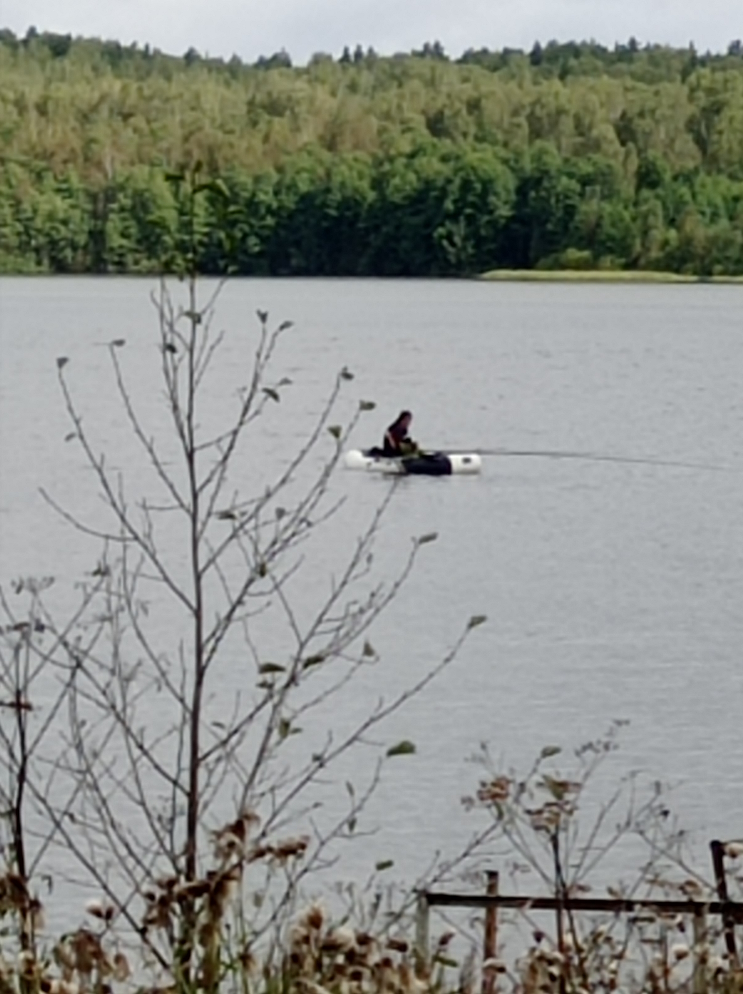 A person is sitting on a white inflatable raft in the middle of a lake. The person is wearing dark clothing and appears to be fishing. The raft is connected to a line that stretches across the water. In the background, there is a dense forest with green trees. The sky is overcast. The foreground is filled with dried plants and branches. The scene is calm and peaceful. The location is a lake in Degtyarsk, Russia.