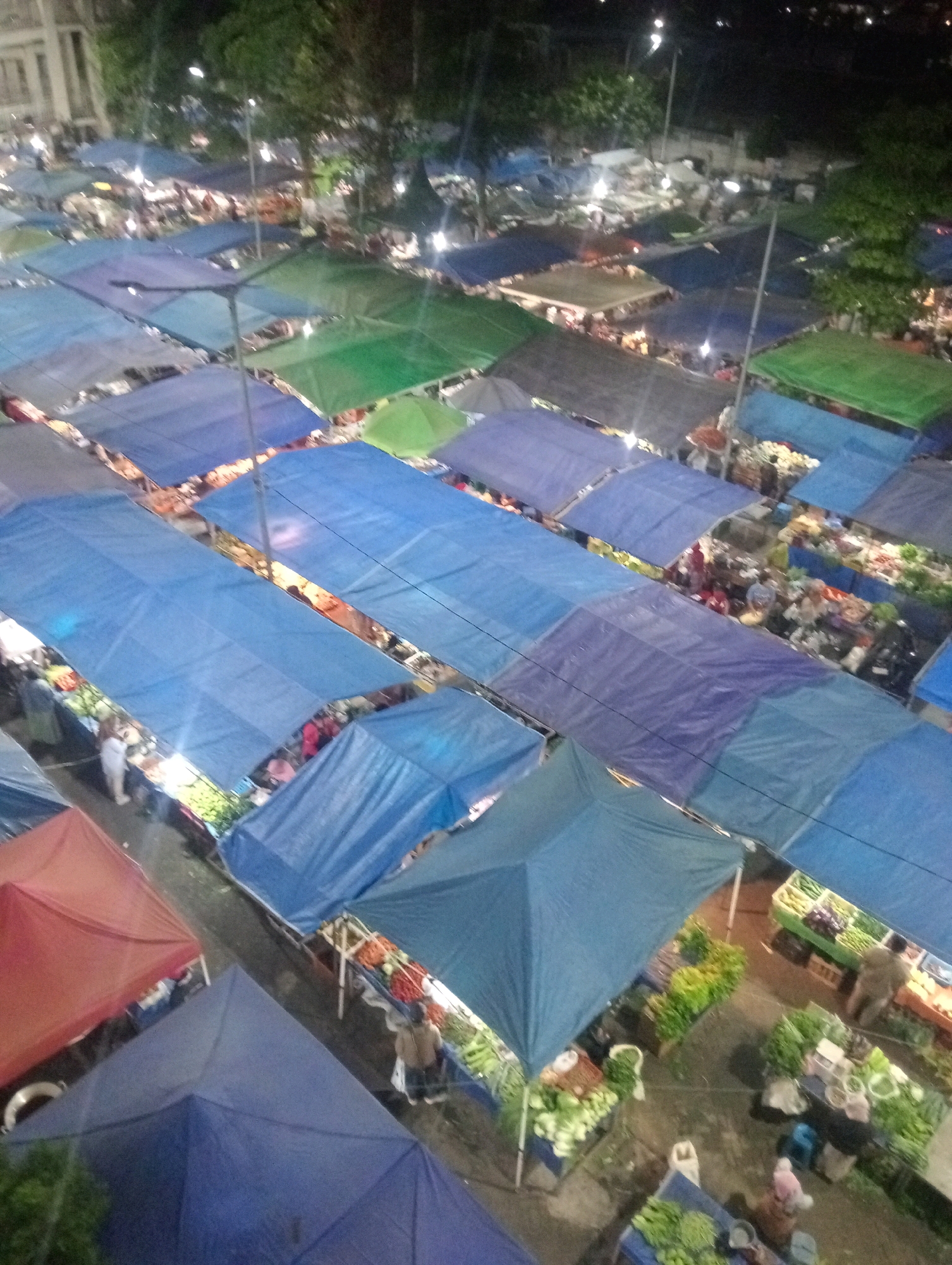 This is an aerial, high-angle, full-shot, low-quality, night-time photograph of a bustling street market in Batu, Indonesia. The market is covered with numerous blue, green, and red tarps, creating a chaotic patchwork of temporary shelters. Stalls are laden with various produce, including what appear to be leafy green vegetables and possibly fruits like tomatoes and eggplants. People are visible milling about, interacting with vendors, and browsing the goods. The lighting is dim, with some artificial lights illuminating the stalls, casting long shadows. The overall impression is one of a lively and active marketplace at night.