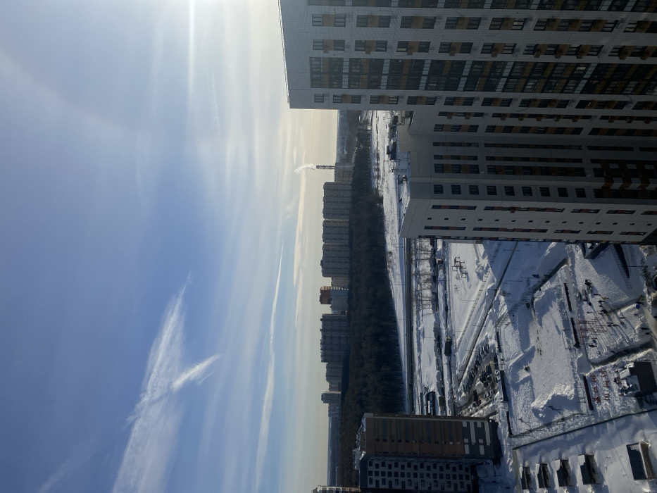 The image captures an expansive, elevated view of a snow-covered urban and forested landscape in Bittsa, Russia, under a clear, bright blue sky with scattered high-altitude clouds. The sun is visible in the upper right, casting a significant glare.
In the immediate foreground, two modern, multi-story residential buildings frame the lower left and right portions of the scene. The building on the right is light-toned with numerous dark-paned windows, while the building on the left is darker in color.
Directly below, a snow-covered ground plane reveals a residential courtyard or recreational area. Approximately 15-20 individuals, visible as small dark figures, are present in various clusters across the snow, suggesting general outdoor presence. The area contains snow-covered structures consistent with playground or outdoor fitness equipment. An adjacent snow-covered parking lot holds several parked vehicles.
The midground features a wide, snow-blanketed expanse, bisected horizontally by a multi-lane road or highway, partially cleared of snow. Beyond the road lies a dense, dark evergreen forest, likely Bittsa Forest, forming a prominent natural barrier.
In the distant background, a dense cluster of multi-story residential buildings extends across the horizon, forming a city skyline. To the far right of this background cityscape, a tall, slender industrial smokestack is visible, emitting a distinct plume of white smoke or steam vertically into the cold air. The overall environment indicates a clear winter day with pervasive snow cover. No specific interactions or detailed actions between individuals are discernible from this distance.