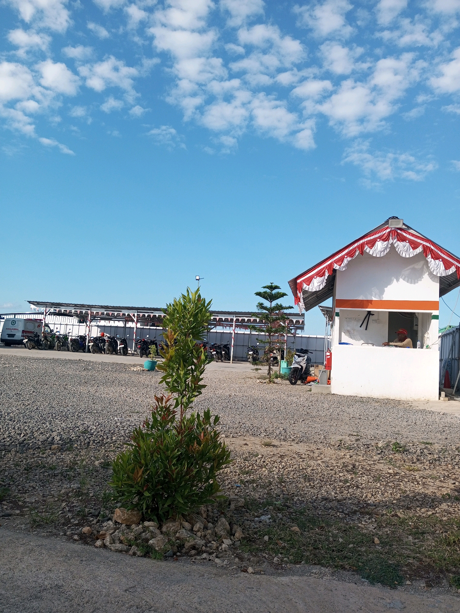 This image shows an outdoor scene with a clear blue sky dotted with white clouds. In the foreground, a small, green shrub with red-tipped leaves grows amidst rocks and gravel. To the right, a small, white building with a red and white draped awning serves as a kiosk or guardhouse. An individual wearing a red cap is visible inside the building. A thin evergreen tree stands to the left of the kiosk. Behind the kiosk, a row of motorcycles is parked under a covered structure with a white roof supported by white posts. A white van with "AMBULANS" written on its side is parked at the far left of the covered area, followed by more motorcycles. The ground is covered in gravel and some patches of grass. The setting appears to be a parking area or a facility in Matasio, Indonesia.