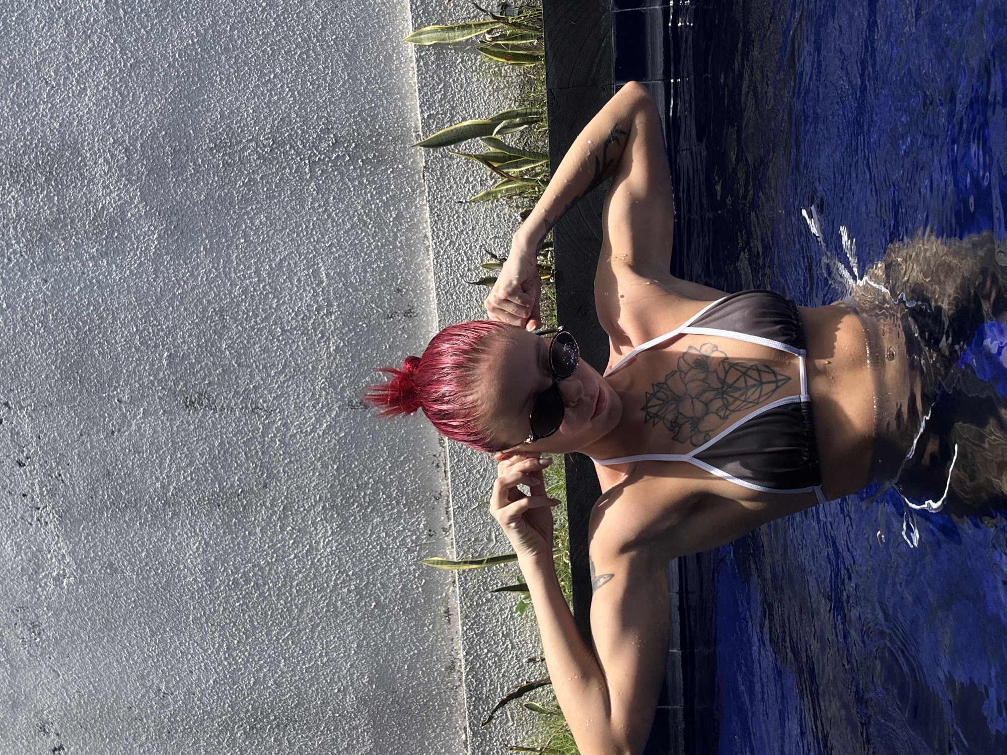 A light-skinned person with red hair is in a swimming pool. The person is wearing a black and white bikini and sunglasses. The person has tattoos on their chest and arms. The swimming pool is located next to a wall with plants growing on top of it. The photo was taken in Dalung, Indonesia.