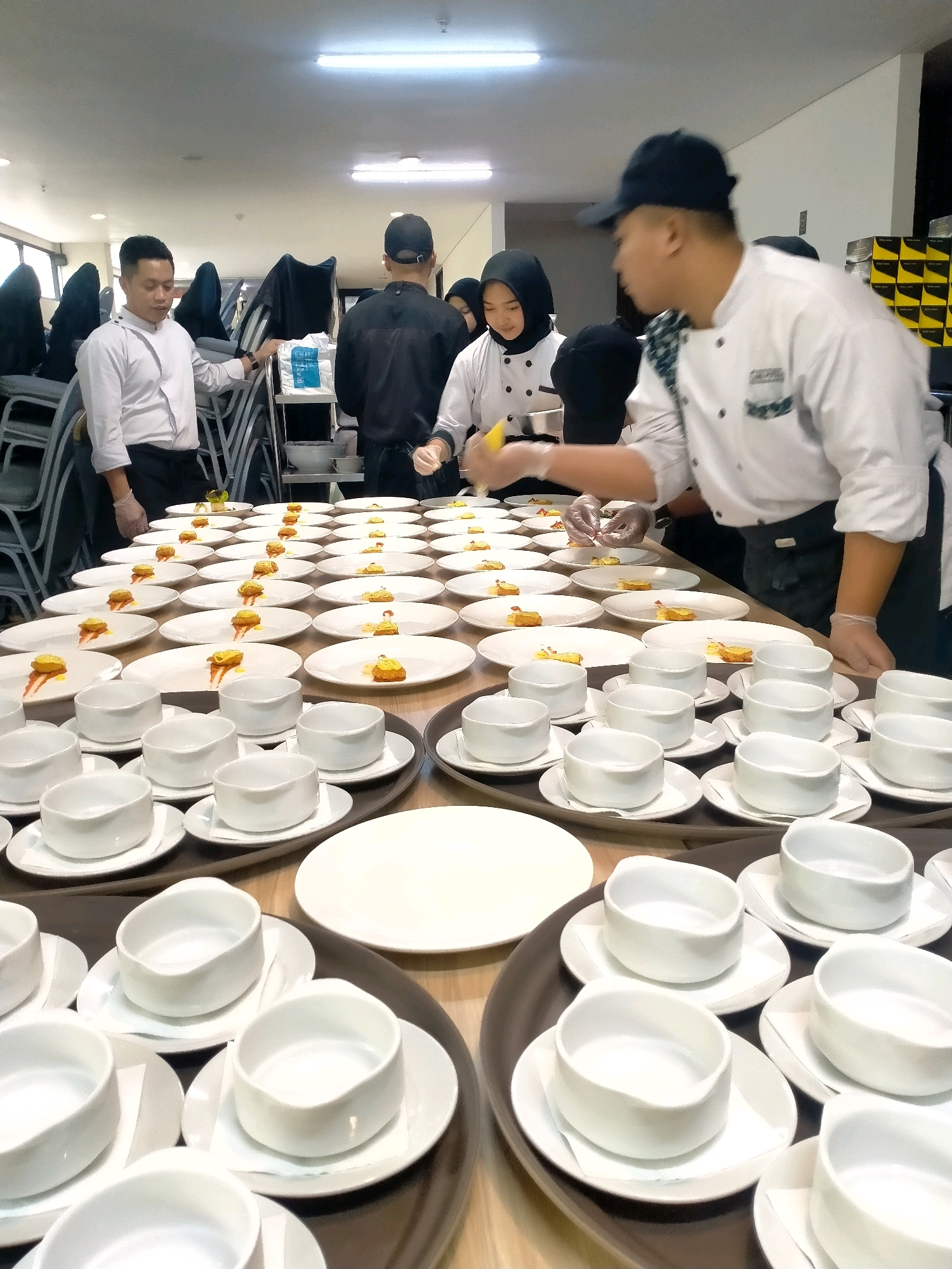 The image depicts a culinary event preparation scene, likely at a hotel or large dining establishment in Batu, Indonesia. The foreground is dominated by a long table covered with numerous white plates and bowls, meticulously arranged for a meal service. Many plates already have small portions of food, some adorned with a dollop of yellow sauce and a garnish, suggesting appetizer or tasting portions. In the mid-ground and background, several individuals, dressed in chef's attire, are actively engaged in plating food. Two individuals are clearly visible in the foreground, one wearing a white chef's jacket and the other a black chef's jacket and cap. A third individual in the background wears a hijab and a chef's jacket, participating in the food preparation. The environment appears to be indoors, possibly a banquet hall or kitchen prep area, with fluorescent lighting overhead and stacks of chairs visible in the background, indicating a large-scale event is being organized. The overall impression is one of organized chaos and precision as the culinary team prepares to serve a meal.