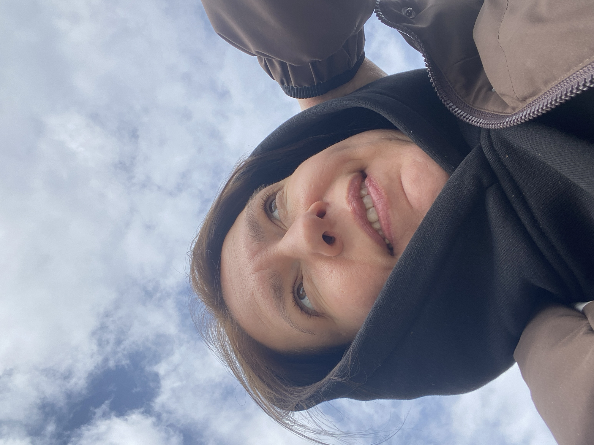 A person is looking down at the camera, smiling. They are wearing a black hoodie and a brown jacket. The background is a blue sky with white clouds. The location is likely outdoors. No evidence exists to indicate this photograph was taken in Chaikovsky, Russia.