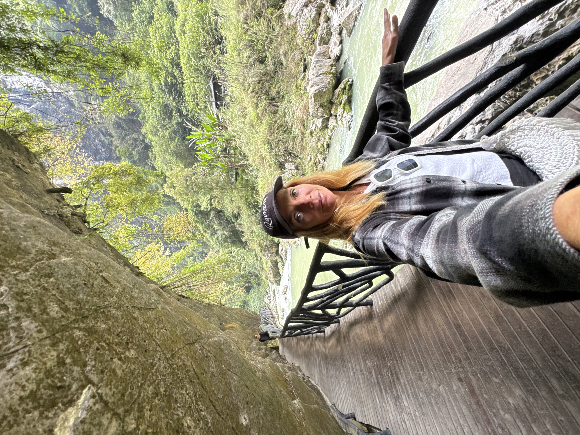 A blonde-haired woman in a grey plaid shirt and baseball cap is taking a selfie on a wooden walkway with a black railing in Zhuoshui, China. The walkway is adjacent to a river flowing through a rocky gorge with lush greenery on the opposite bank. The camera angle is tilted, emphasizing the steepness of the terrain. A man in black can be seen further down the walkway.