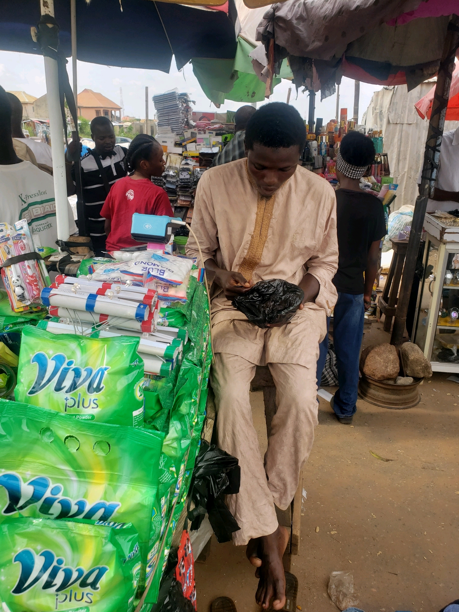 The image depicts a bustling outdoor market scene in Sabon Tasha, Nigeria. A young man in a light-colored tunic sits on a wooden stool, engrossed in a black plastic bag he is holding. To his left, a stall is laden with green packaging of "Viva plus" powder, along with other items like toothbrushes and personal care products. Behind him, other individuals are visible, including a person wearing a red shirt and another in a black shirt and blue jeans. The market appears to be under a makeshift canopy, with corrugated metal structures and colorful fabrics forming the overhead cover. The ground is dusty and uneven, indicative of a typical Nigerian market environment. Various goods, including bottles of what appear to be perfumes or lotions, are displayed on shelves. The overall atmosphere suggests active commerce in a vibrant community setting.
