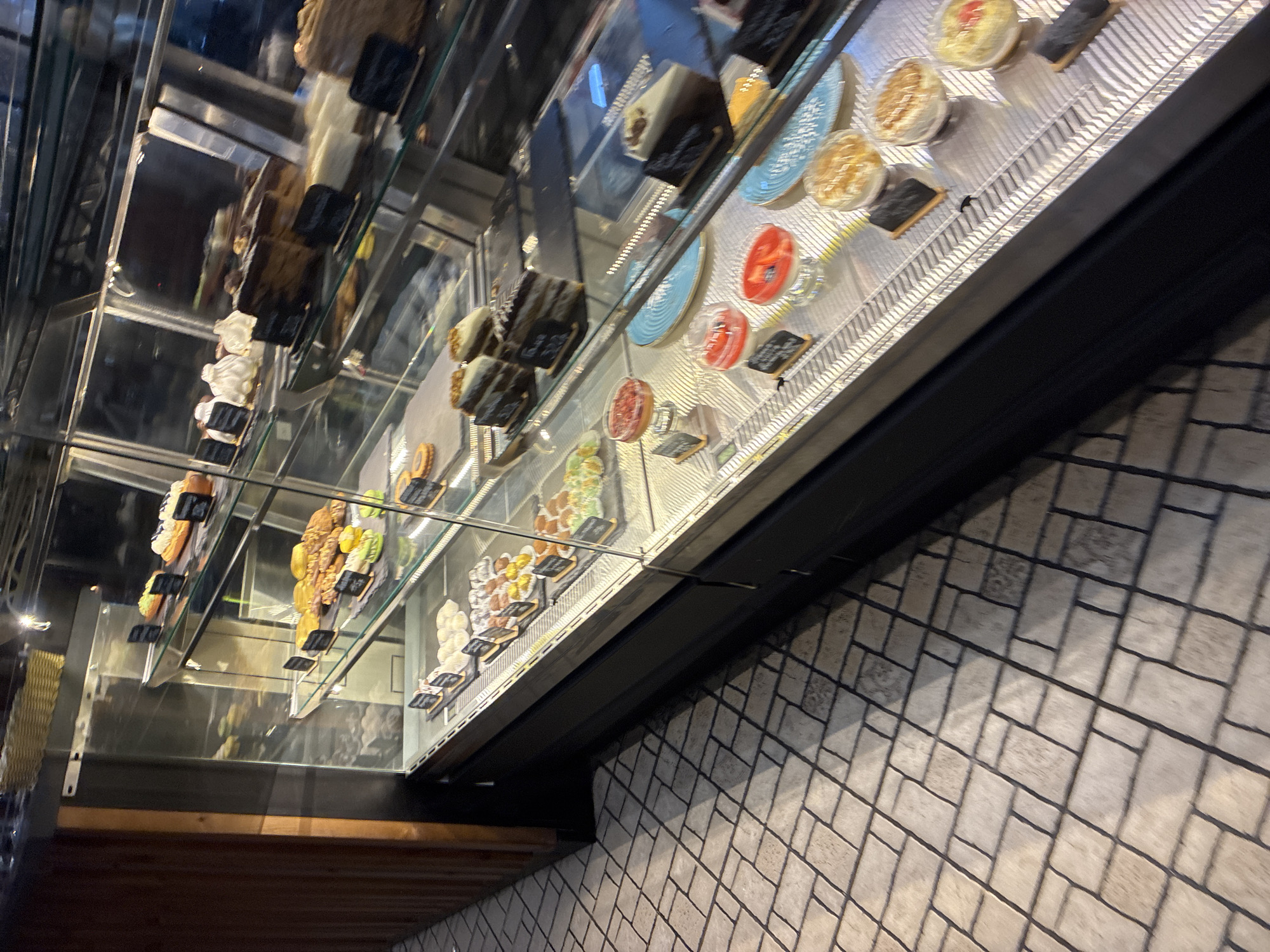 A display case filled with various pastries and desserts is shown in this photograph. The display case is glass-fronted and appears to be in a bakery or dessert shop. The pastries are arranged on multiple shelves. On the top shelf, there are several small, round pastries topped with what appears to be whipped cream and fruit. Below that, a variety of cakes and tarts are visible, some decorated with intricate icing designs. Further down, individual dessert cups containing layered creams and fruits are displayed. The floor in front of the display case is tiled with a pattern of light-colored rectangular and square tiles. The background is dark, suggesting an indoor setting with low lighting.