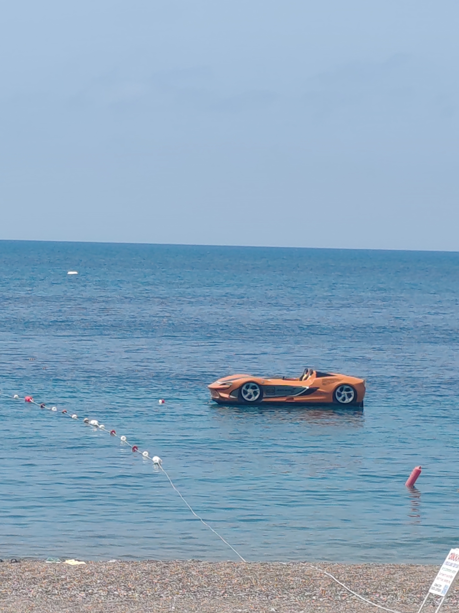 The image depicts a beach scene in Alanya, Turkey, featuring a bright orange sports car-shaped paddle boat floating in the calm, blue sea. A line of buoys with red and white markings is visible on the left side of the frame, marking a swimming area. Another solitary red buoy is positioned closer to the shore on the right. The beach itself is composed of small pebbles. The sky is clear and pale blue, blending with the sea along the horizon. A small white object is also floating in the water at a distance. A white sign with lettering is placed on the shore on the right side of the frame.