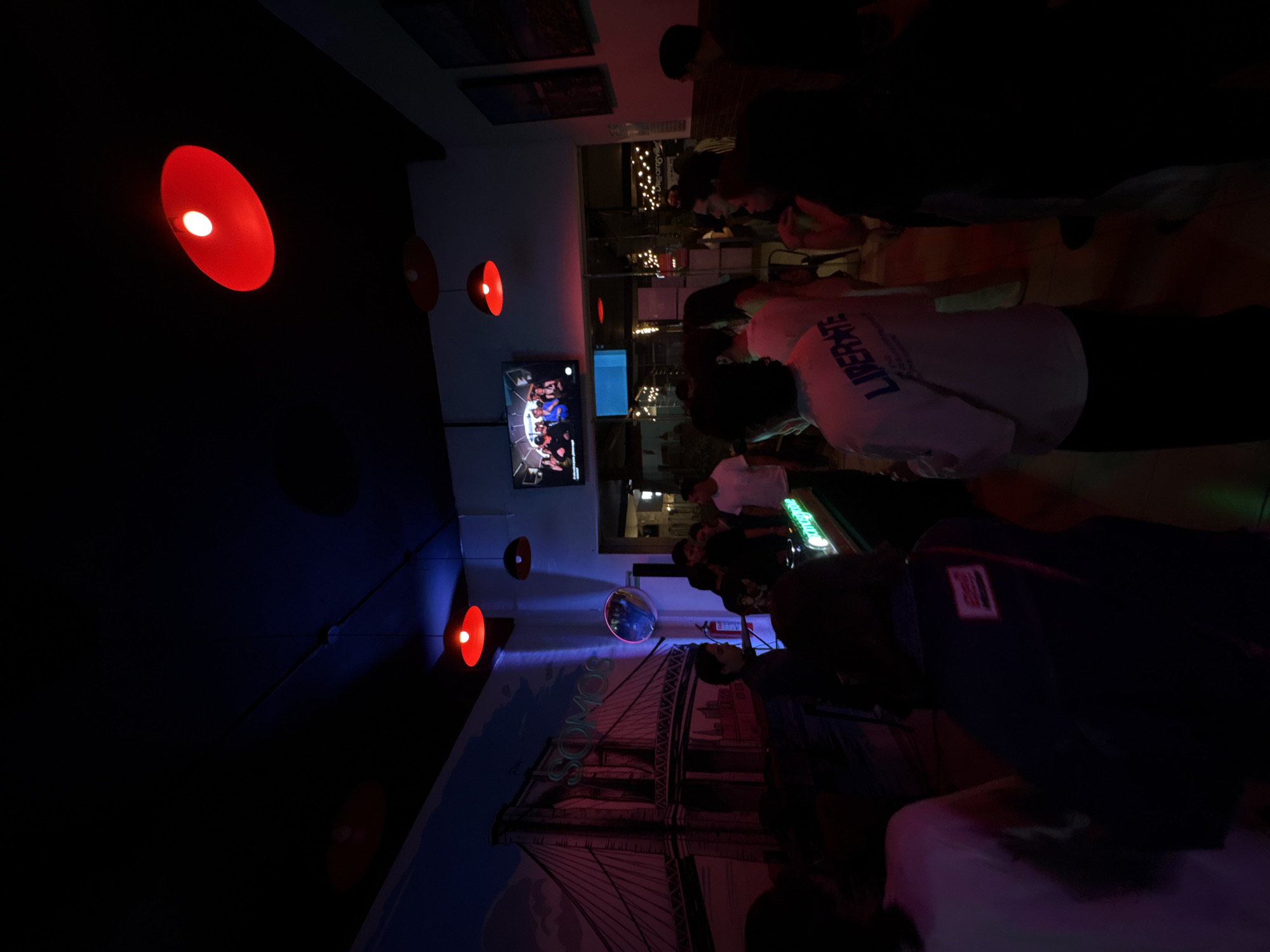 The image depicts a dimly lit indoor space, likely a bar or restaurant in San Bernardino Tlaxcalancingo, Mexico. Red pendant lights are suspended from the ceiling, casting a warm glow. A television screen displays an image of people, possibly a sporting event or a show. Patrons are gathered, some standing and others seated, interacting with each other. In the background, there is a large mural depicting the Brooklyn Bridge. The lighting and the mural together create a casual atmosphere.
