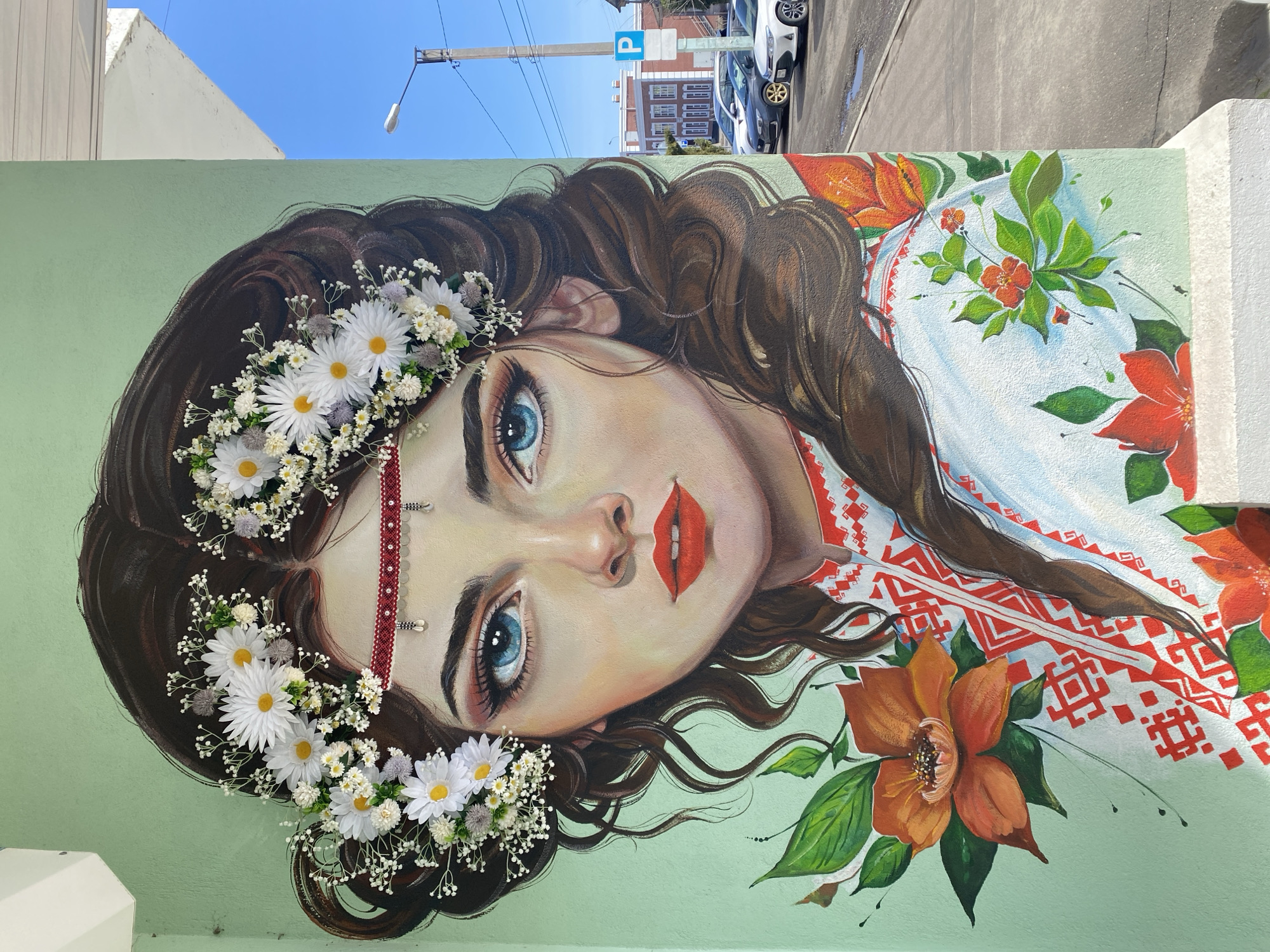 A mural on a light green wall depicts a woman with light skin, blue eyes, and brown wavy hair. She wears a crown of white daisies and baby's breath, along with a red beaded headpiece. Her lips are painted red. Her dress is white with traditional red geometric patterns, and includes orange flowers and green leaves. The painting appears to be outdoors in Yoshkar-Ola, Russia. This is deduced from the parking sign with the letter "P" and the architectural style of the building visible in the background, which are consistent with Russian urban landscapes. Cars are parked nearby. Overhead power lines cross the sky.