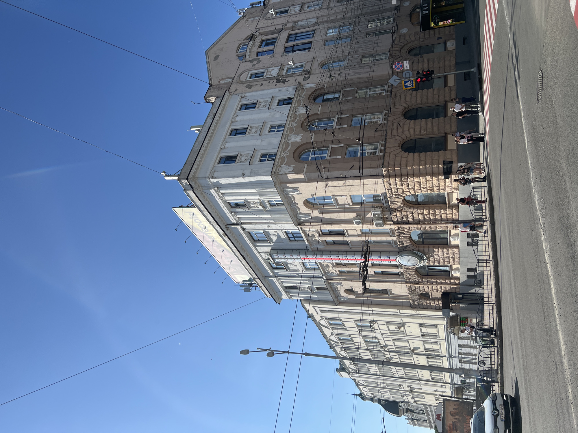 The image shows a street scene in Kharkiv, Ukraine, featuring a large building with classical architectural elements and a prominent thermometer on its facade. The building has a light pink and white exterior, with multiple stories of windows. A large round clock is visible on the lower portion of the building. There are several people standing on the sidewalk and at a crosswalk. Traffic lights are visible, and a white car is driving on the street. Overhead wires crisscross the sky, suggesting an urban environment with public transportation, likely trams. The sky is clear and blue. The building appears to be located at the intersection of a street and a wide avenue, with a pedestrian crossing. There is signage on the building, some of which is in Cyrillic script. The overall impression is of a sunny day in a bustling city.