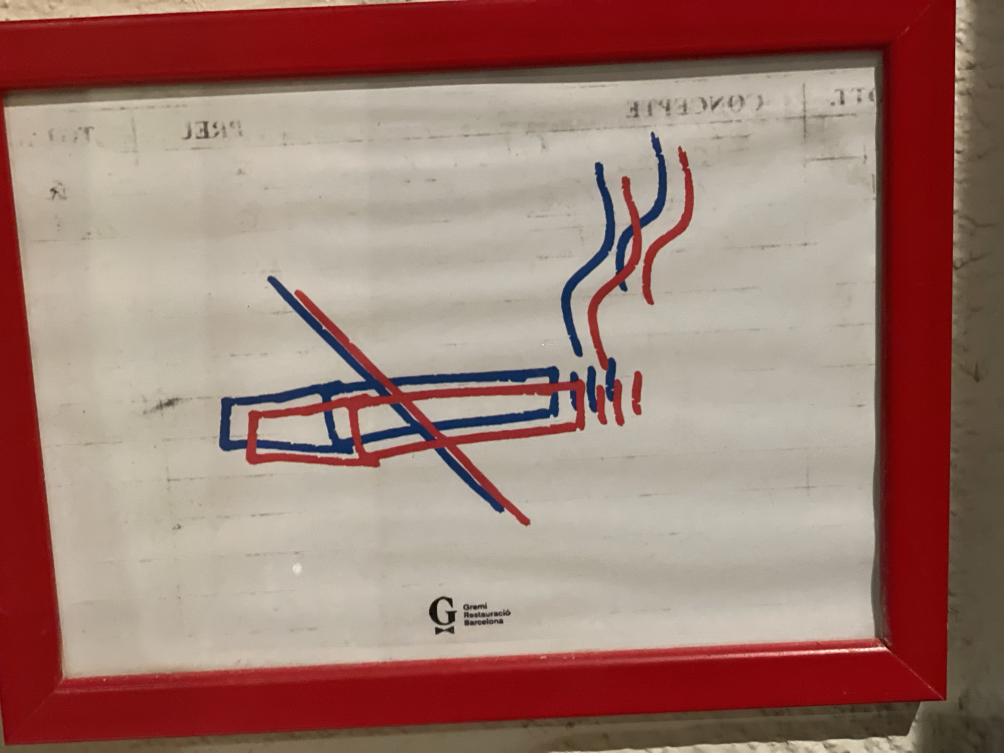 A rectangular sign, enclosed in a bright red frame, is mounted on a light-colored, textured wall. The sign features a white background with faint, reversed, and partially obscured text visible at the top, including segments such as "LE", "CONCEBIE", "BBEI", and "T. J". The central element of the sign is a hand-drawn illustration depicting two stylized cigarettes. One cigarette, rendered in blue outline, lies horizontally. Adjacent to its right end, two wavy blue lines ascend, representing smoke. Overlapping this, a second cigarette, rendered in red outline, also lies horizontally, slightly below the blue one, with two wavy red lines ascending from its right end, representing smoke. Both cigarettes are diagonally crossed by a thick blue line and a thick red line, originating from the upper left and extending to the lower right, signifying prohibition. In the lower left quadrant of the sign, a black logo is present, featuring a stylized capital "G" alongside the vertically stacked text: "Gremi", "Restauració", and "Barcelona". This logo identifies the sign as originating from the Restaurant Guild of Barcelona, unequivocally placing the scene within the city of Barcelona, Spain.