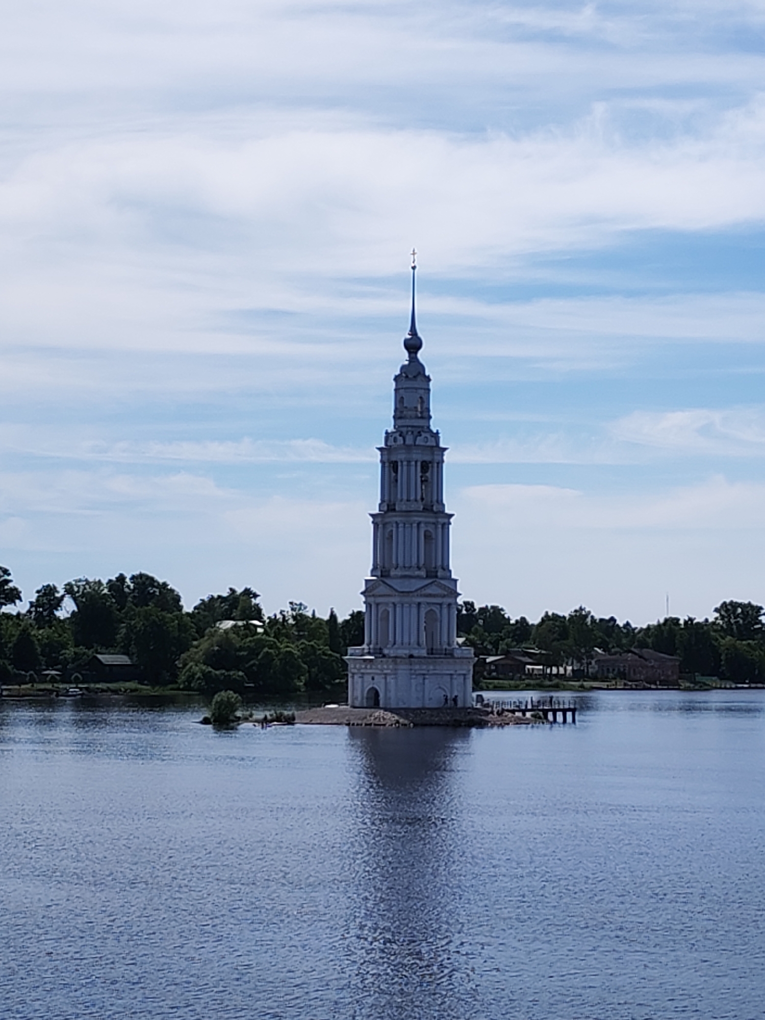 The image displays the flooded bell tower of St. Nicholas Cathedral (Nikolsky Cathedral) in Kalyazin, Tver Oblast, Russia, situated centrally within a wide body of water. The bell tower is a tall, multi-tiered structure of light-colored material, featuring classical architectural elements including columns and arched openings on its upper levels. It is topped by a dark spire and an Orthodox cross. The base of the bell tower rests on a small, partially submerged rocky island. A short wooden pier extends from the bell tower's base towards the right. Several small human figures are visible on this pier and near the tower's immediate foundation, indicating visitation. The surrounding water shows ripples and reflects the bell tower faintly. The background on both sides of the waterway features dense green tree lines. On the left bank, scattered buildings are partially obscured by the foliage. On the right bank, more trees are visible, along with a larger building with arched windows. The sky above is light blue with elongated, scattered white clouds.