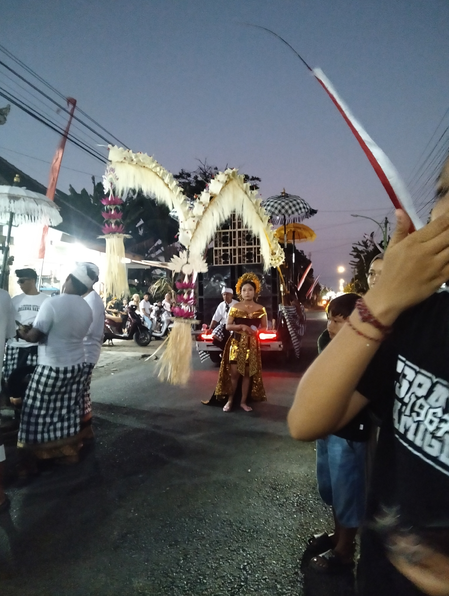 The image depicts an outdoor scene at dusk in Sumbersewu, Indonesia, likely during a cultural event or parade. In the foreground, a person is holding up a red and white striped banner. The central subject is a young woman in traditional Balinese attire, adorned in a gold ornate dress and headdress, standing on a street. Behind her, a truck is decorated with elaborate white and gold streamers and a black and white checkered umbrella. Several people are visible in the background, some on motorcycles and others standing around. The street is wet, reflecting the surrounding lights. The atmosphere suggests a festive or ceremonial gathering taking place at twilight.