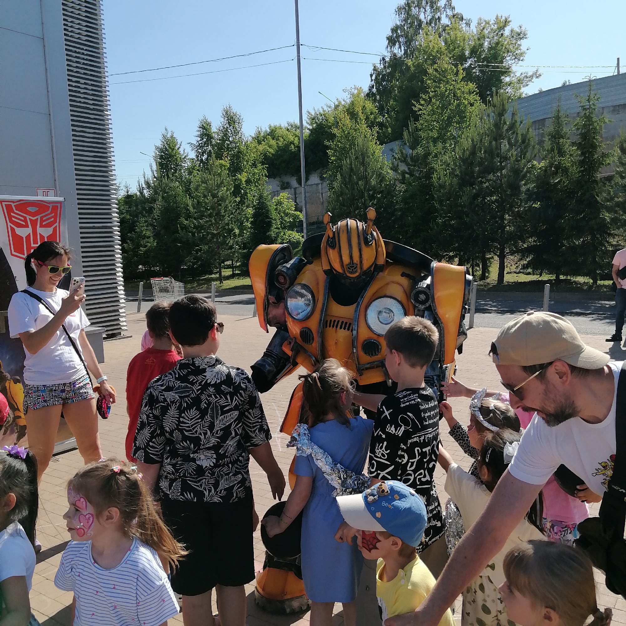 The image depicts an outdoor gathering in Трубачеевка, Russia, under clear blue skies. The central focus is a person inside a yellow and black Bumblebee costume, resembling a Volkswagen Beetle, with functional-looking headlights and a stylized head. Several children and adults are congregated around the costume.
On the left side of the frame, a woman with dark hair tied in a ponytail wears a white t-shirt, patterned shorts, and yellow-green framed sunglasses. She smiles while looking at and holding a white smartphone, seemingly capturing a photo or video. A black crossbody bag is visible, and she holds a colorful object in her right hand. Behind her and to the left, a large, grey building structure with vertical ribbed paneling is visible. A prominent red and white Autobot logo (Transformers symbol) is affixed to this structure, with small red "ПГ" or similar characters below it.
Children of various ages surround the Bumblebee costume. One girl in the foreground wears a white striped t-shirt and has pink heart-shaped face paint on her left cheek. Another small boy on the bottom right wears a blue and white cap with a bear motif and a yellow t-shirt, featuring red spiderweb face paint on his right cheek. A girl in a blue dress with silver foil material draped over her shoulders stands near the costume, as do two boys seen from behind, one in a black and white leaf-patterned shirt and sunglasses, and another in a black t-shirt with white script. Some children appear to be touching the costume.
On the right side, an adult male with a beard wears a light-colored cap and sunglasses, a white t-shirt, and a dark camera strap over his shoulder. He looks downwards. In the middle ground, lush green trees and foliage dominate the background, along with a grey utility pole and overhead wires. A distant building with a grey roof is visible through the trees. Further to the right, a white shopping cart is parked near a paved area. The ground where the people are standing is paved with light-colored rectangular blocks. The scene is brightly illuminated by direct sunlight.