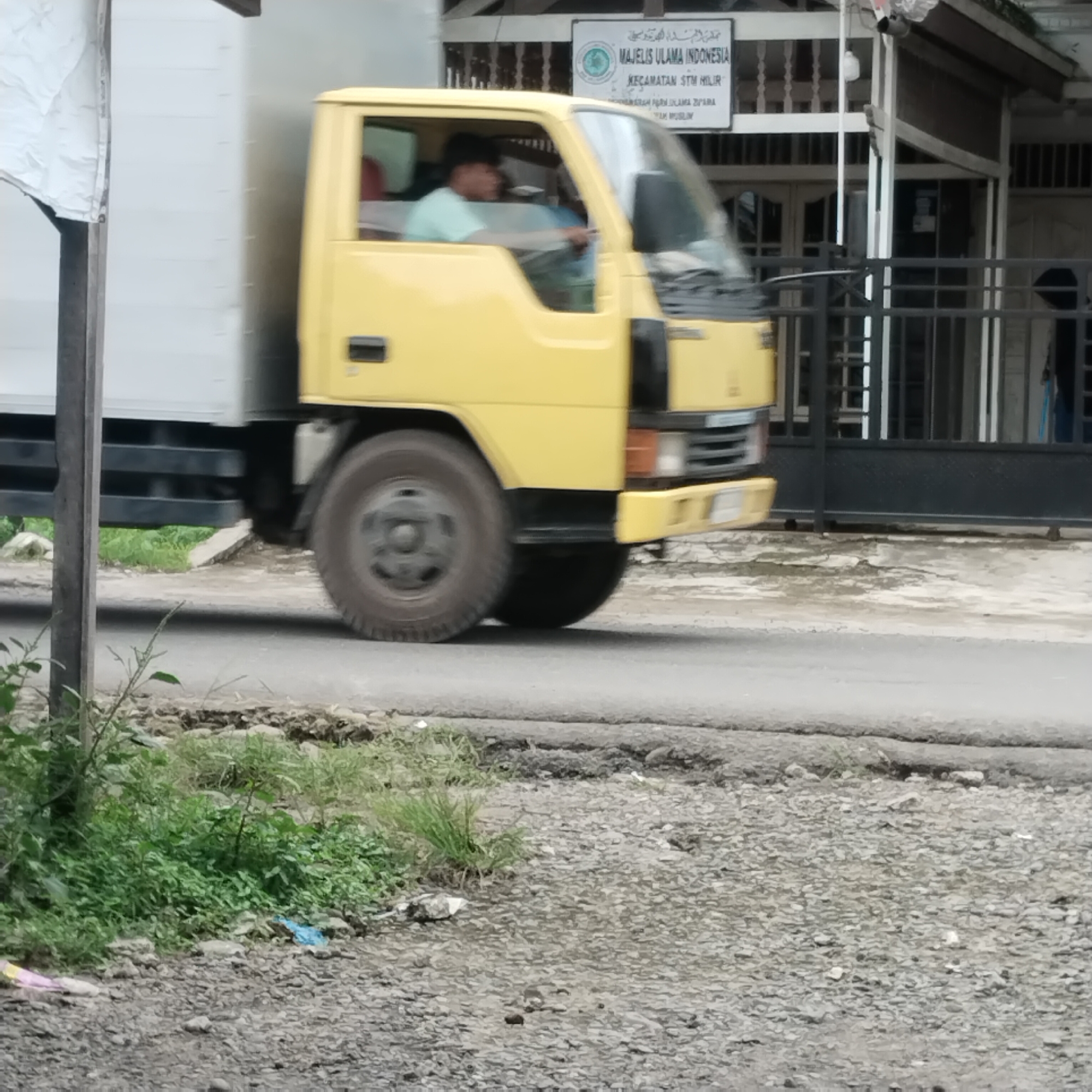 In Talun Kenas, Indonesia, a yellow truck is captured in motion on a paved road. The truck's driver, a male with dark hair, is visible behind the windshield, wearing a light blue shirt and gripping the steering wheel. The truck is in the foreground, with its front wheel prominently displayed. To the left of the truck, a white tarp is attached to a wooden post, and beyond that, the side of a white trailer is visible. Behind the truck, a building with a black metal fence and open windows is present. A white sign with Arabic and Latin script is affixed to the building. The road is lined with green vegetation and gravel. The overall scene depicts a common street view with a moving vehicle.