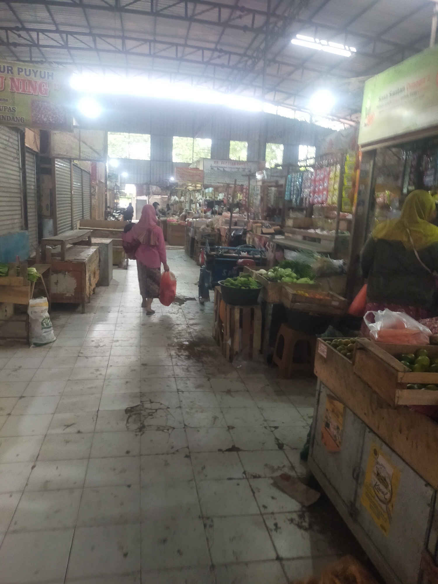 The image depicts an indoor market scene, likely in Trayeman, Indonesia, characterized by its tiled floor, metal support structures, and corrugated metal roof with large windows allowing diffused light. In the foreground, a woman wearing a pink headscarf and traditional clothing walks away from the camera, carrying two plastic bags. To her right, a stall displays various vegetables, including cucumbers and carrots, in wooden crates. Further down the aisle, other market stalls are visible, some with closed shutters and others displaying goods such as packaged food items and produce. The overall atmosphere suggests a bustling, everyday market environment.