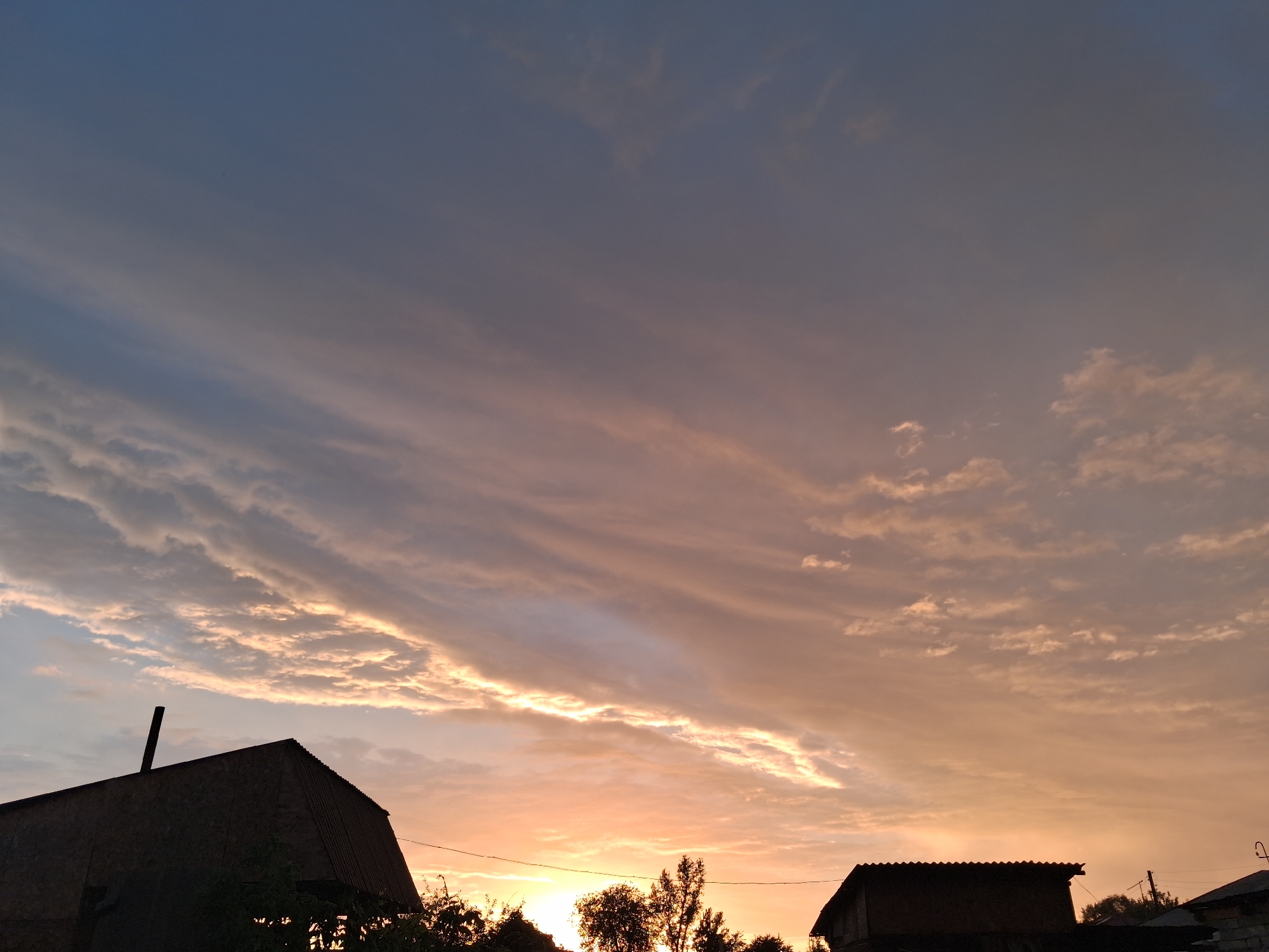 The image captures a twilight sky over the city of Semey, Kazakhstan. The sky is a gradient of deep blue at the top, transitioning to hues of pink, orange, and yellow near the horizon, illuminated by the setting sun. Wispy, elongated clouds streak across the sky, catching the warm light. In the foreground, silhouetted against the vibrant sky, are the rooftops of several buildings. The roof of a large, gabled structure with a prominent chimney dominates the left side of the frame. To the right, another, smaller building with a dark, sloped roof is partially visible. Trees and foliage are also silhouetted along the bottom edge of the image, suggesting a suburban or rural setting within the city. The scene is devoid of people or any explicit activity.