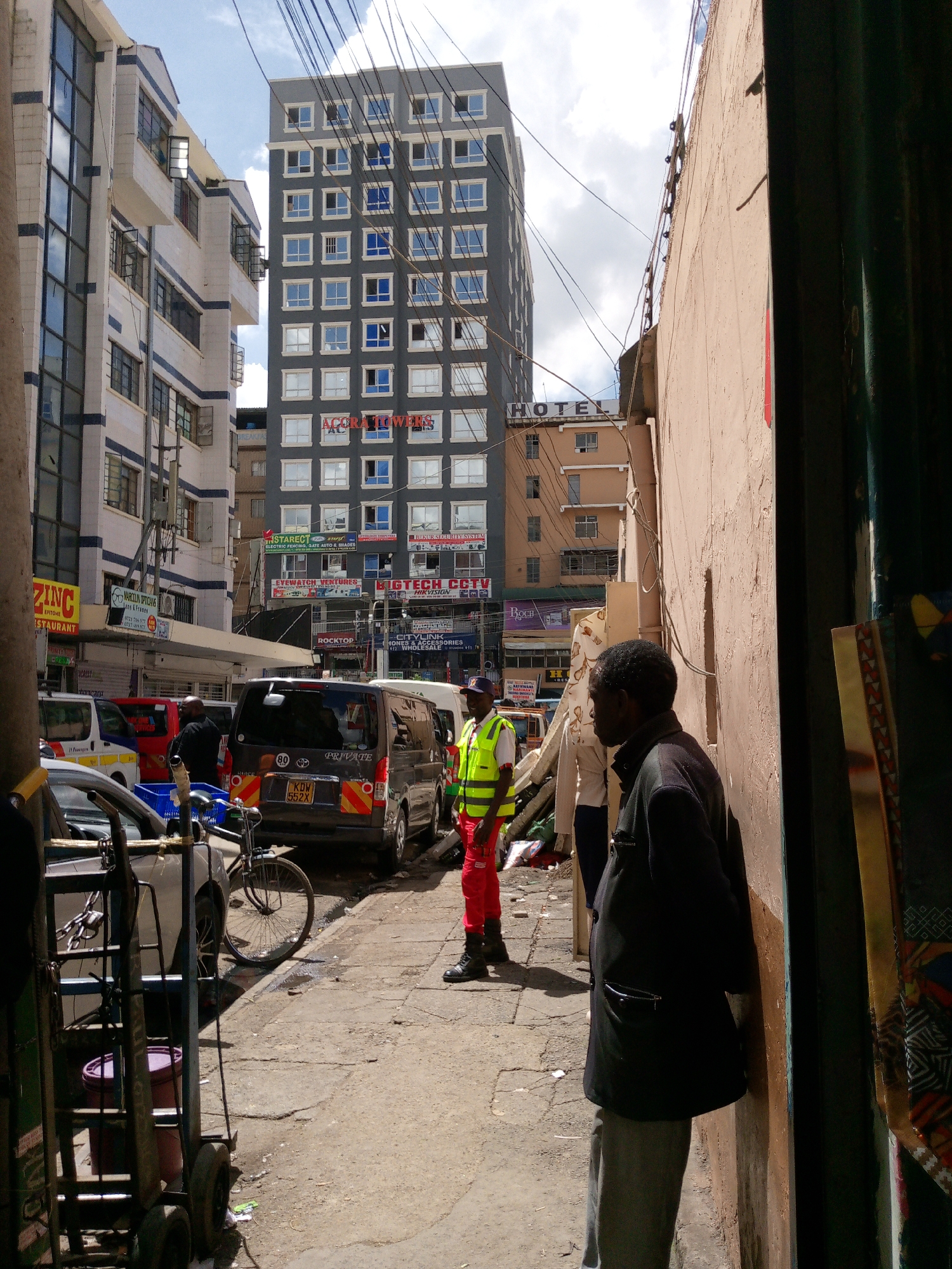 The image captures a daytime street scene in Nairobi, Kenya, specifically identifying the prominent "ACORA TOWERS" in the background, likely placing the location within the city's Central Business District near areas like Biashara Street or Luthuli Avenue. The perspective is from a narrow thoroughfare or alleyway, looking towards a busier street.
In the immediate foreground on the right, a dark-skinned male with short, dark, kinky hair, dressed in a dark jacket and light grey trousers, leans against a light brown, weathered wall. His right hand is positioned near his mouth or chin. Directly to his left, slightly further back, stands a dark-skinned male wearing a bright yellow high-visibility vest over a dark garment, red trousers, and dark boots, with a dark cap on his head. He stands upright, facing generally forward.
Behind these two individuals, on the street, a dark-colored minivan (license plate KDW 552X) with a rear speed limit sticker of "80" and red/yellow reflective tape is either parked or moving slowly. The word "PRIVATE" is visible on its tailgate. To the left of the minivan, the front portion of a silver/grey car is visible, next to which a blue plastic crate rests on the sidewalk. A black bicycle is leaning against this silver car. A metal hand-cart or trolley, with a dark red bucket near its base, occupies the far left foreground.
The street surface is uneven, worn concrete or asphalt, with visible debris. Numerous overhead electrical wires crisscross the sky.
The background is dominated by multi-story buildings under a partly cloudy sky. The tallest is "ACORA TOWERS," a modern-looking grey building with many square windows, some exhibiting a blue tint. Its lower facade is covered with multiple commercial signs, including "STAREX ELECTRIC FENCING," "EYEWATCH VENTURES," and "BIGTECH CCTV." To the left of Acora Towers, another multi-story building features a white exterior with blue horizontal stripes and large windows, displaying signs for "ZINC RESTAURANT" and "MAROON OPTICIANS." To the right of Acora Towers, behind the foreground wall, a light brown building with the word "HOTEL" prominently displayed on its roofline is visible.