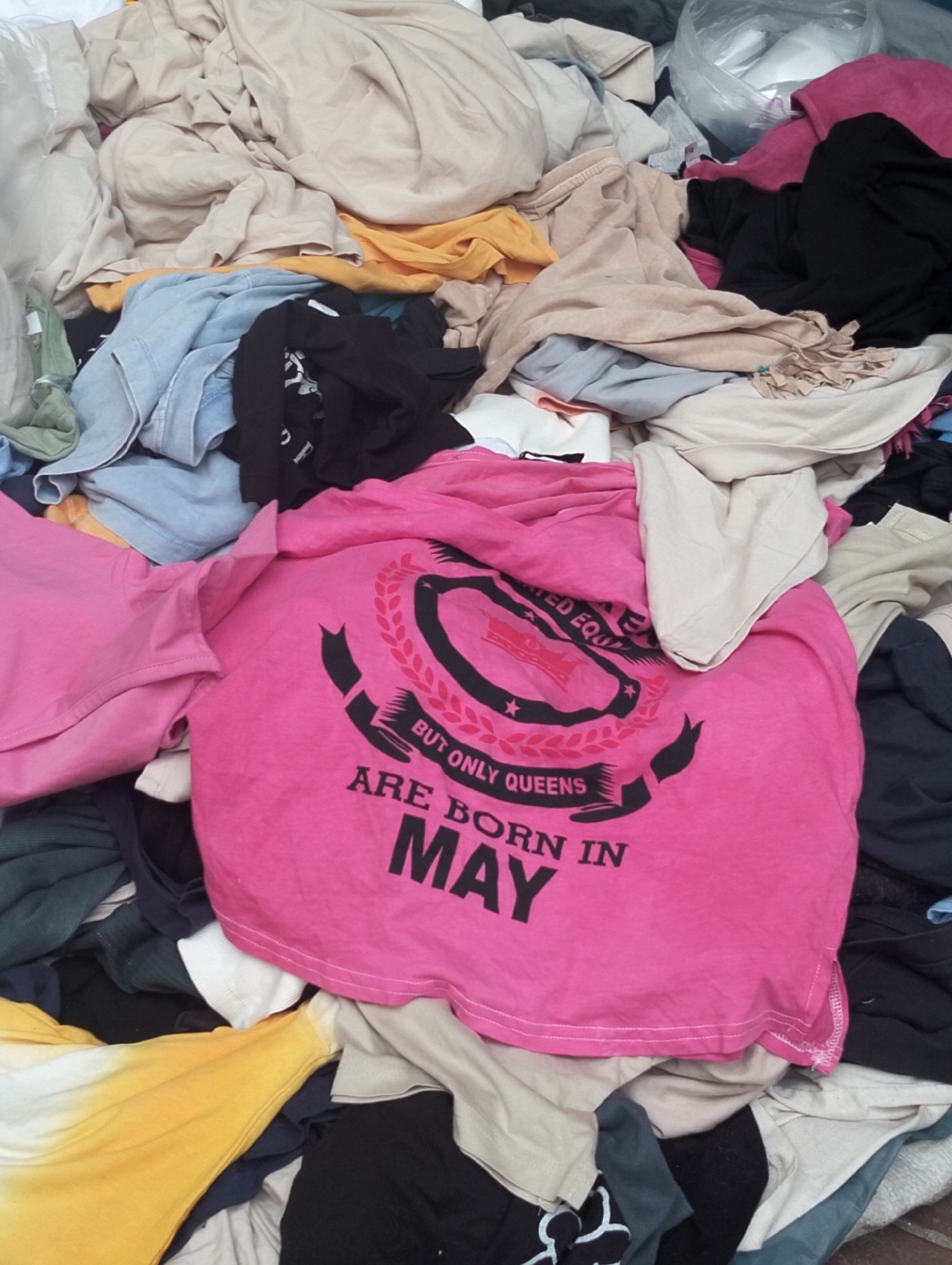 The image depicts a pile of discarded clothing. A pink t-shirt with the text "BUT ONLY QUEENS ARE BORN IN MAY" is prominently visible on top. Other garments in various colors and textures are scattered throughout the pile. The location is unknown.