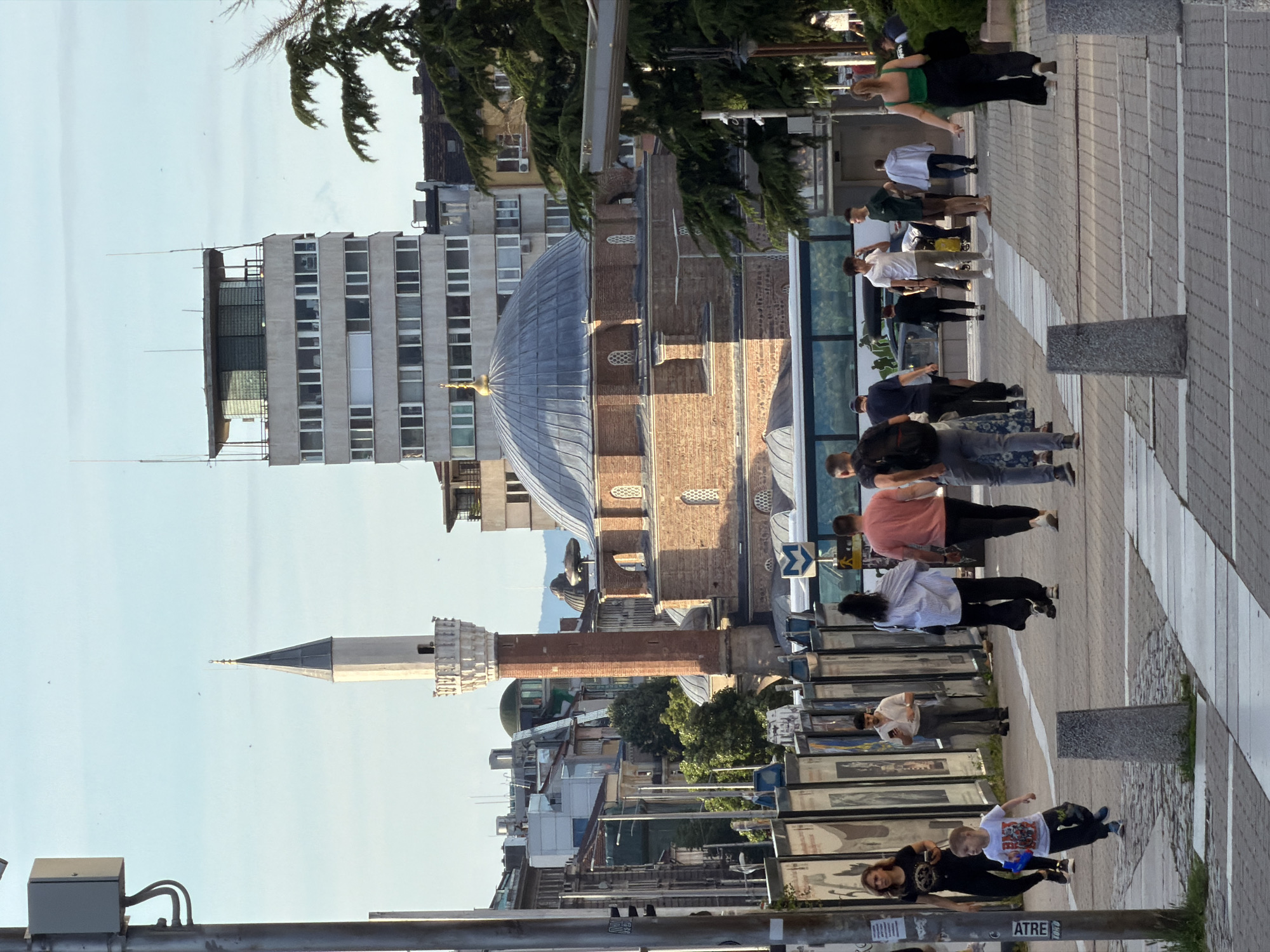 The image shows the Banya Bashi Mosque and the adjacent city landscape in Sofia, Bulgaria, with a group of people walking on a wide sidewalk adjacent to an urban thoroughfare. The mosque is the most prominent architectural element, featuring a large dome and a minaret. The surrounding buildings are a mix of modern and older architecture. On the sidewalk, people are walking in different directions and their clothing is casual.
