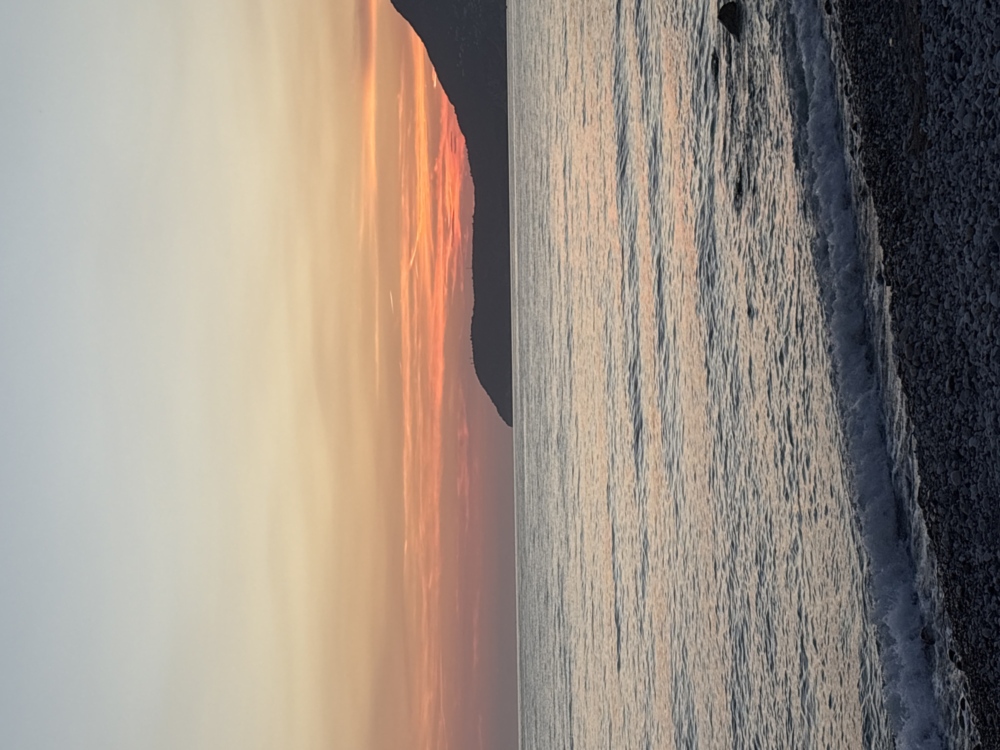 The image depicts a tranquil seascape at sunset, as observed from the pebbled shore of Bar, Montenegro. The horizon is marked by a gentle gradient of colors in the sky, transitioning from a pale blue at the top to warm hues of orange and pink near the horizon, indicating the setting sun. A dark, silhouetted mountain range forms the backdrop, its outline sharp against the vibrant sky. The sea is calm, with subtle ripples reflecting the colors of the sunset. Small waves are breaking softly on the shore, which is composed of numerous dark and light-colored pebbles. No people or specific objects of interest are visible in the frame, creating a serene and natural landscape.