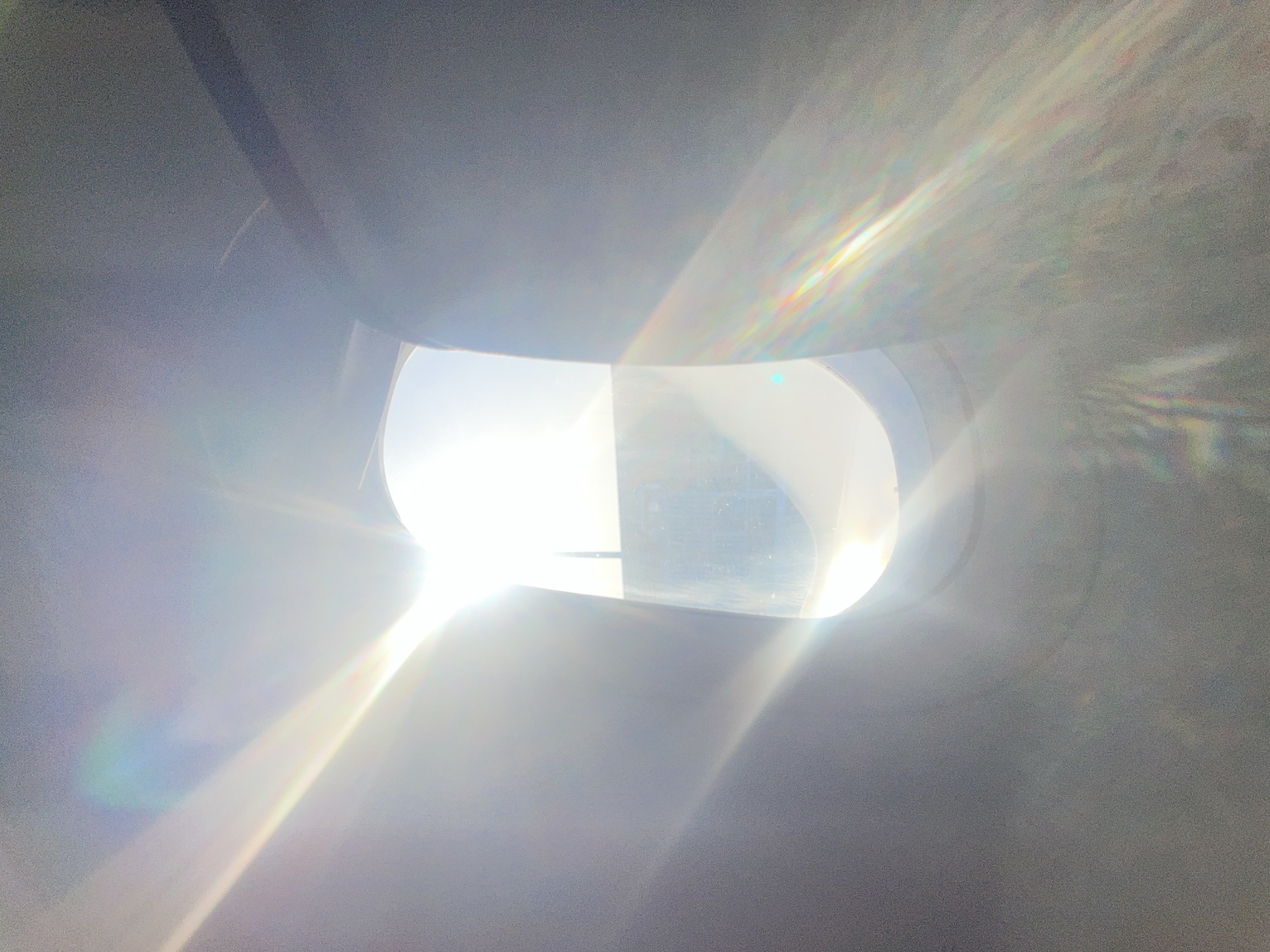 The image shows a bright, sunlit view from inside a structure. The primary focal point is a window, shaped irregularly, through which intense sunlight streams. Outside the window, hints of buildings and a hazy skyline are visible, suggesting an urban or semi-urban environment. The interior of the structure is predominantly gray, with light reflecting off surfaces, creating glare and obscuring details. The scene overall is characterized by strong contrast between light and shadow.