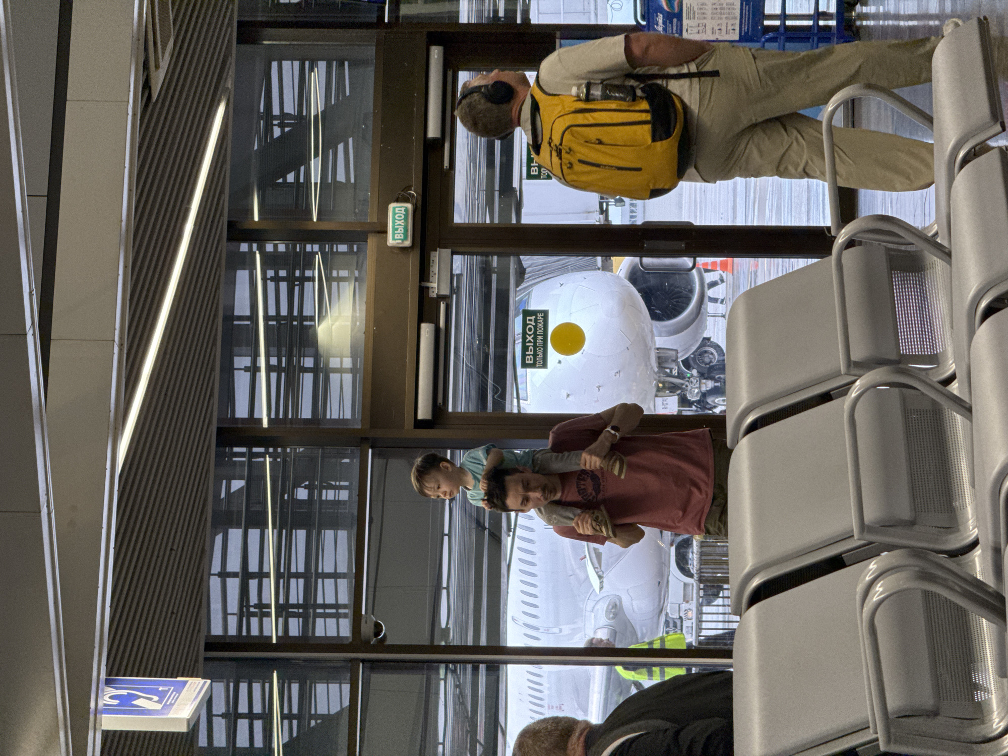 The image depicts the interior of an airport terminal in Sochi, Russia. A man wearing a yellow backpack and khaki pants stands near a door. A father carrying a child on his shoulders looks out the window, where an airplane is visible. Seating is also present in the foreground of the photo.