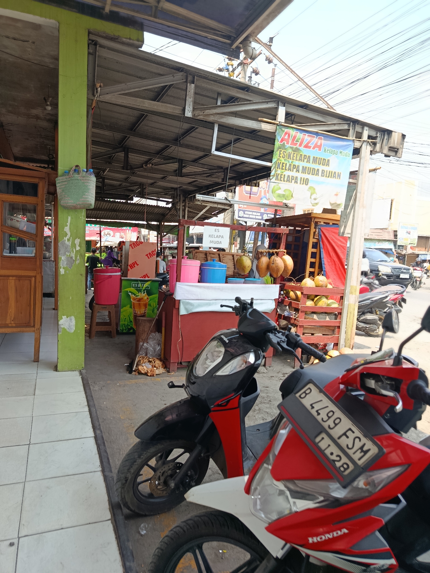 The image shows an outdoor scene in the city of Wanasari, Indonesia, featuring a small food and drink stall specializing in young coconut. Two motorbikes are parked in the foreground, with the closest one being red and white and the second one black and red. The stall itself is partially covered by a corrugated metal roof supported by wooden beams. A green pole stands to the left, with a woven basket holding bottles hanging from it. The stall displays coconuts in wooden crates and a sign in Indonesian that reads "ALIZA ES KELAPA MUDA" which translates to "ALIZA YOUNG COCONUT ICE," advertising various coconut-based drinks. Behind the stall, a black SUV is visible, and further down the street, other vehicles and buildings suggest a commercial area. The ground appears to be a mix of sand and concrete. The lighting indicates it is daytime, likely sunny.
