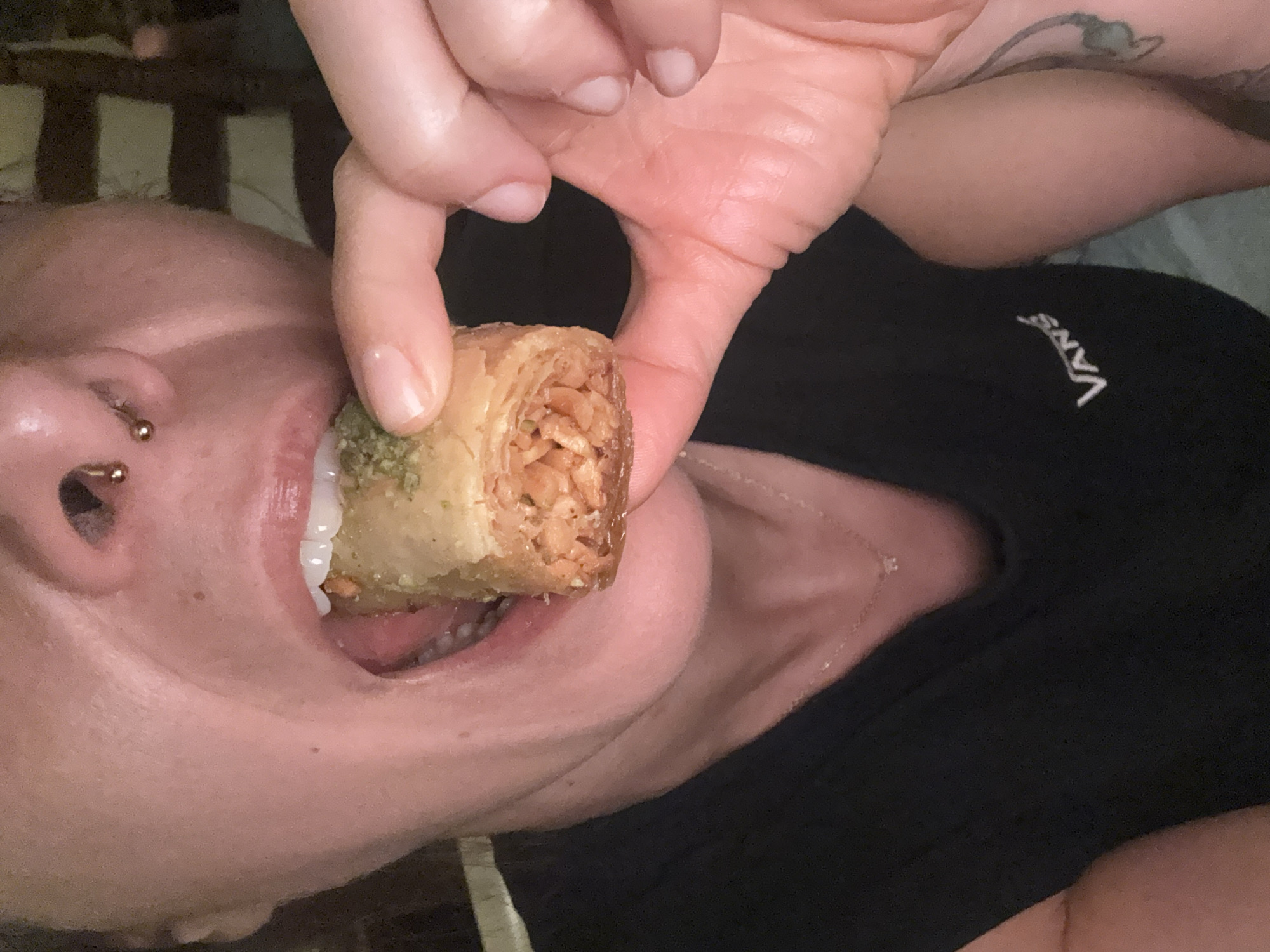 A close-up, horizontally oriented shot captures a person about to eat a piece of baklava. The person's mouth is open, with their lips slightly parted, revealing teeth and tongue. A golden-brown piece of baklava, dusted with green chopped nuts, is held between their fingers, positioned near the person's mouth. The baklava appears layered and flaky, with visible chopped nut filling.
The person has a nose ring on their left nostril. Their skin is visible in the foreground, with some slight imperfections and freckles. The background is dark and out of focus, suggesting an indoor setting. Part of the person's torso is visible, wearing a black t-shirt with the white Vans logo embroidered on it. There's a hint of a tattoo on their upper right arm.
The lighting appears to be artificial and warm, casting subtle shadows and highlighting the texture of the skin and the baklava. The overall mood is casual and intimate, focusing on the act of enjoying a sweet treat. The image does not provide specific cues about the time of day or weather. The location context of Barcelona, Spain, is provided separately and is not directly evident from visual cues within the image.