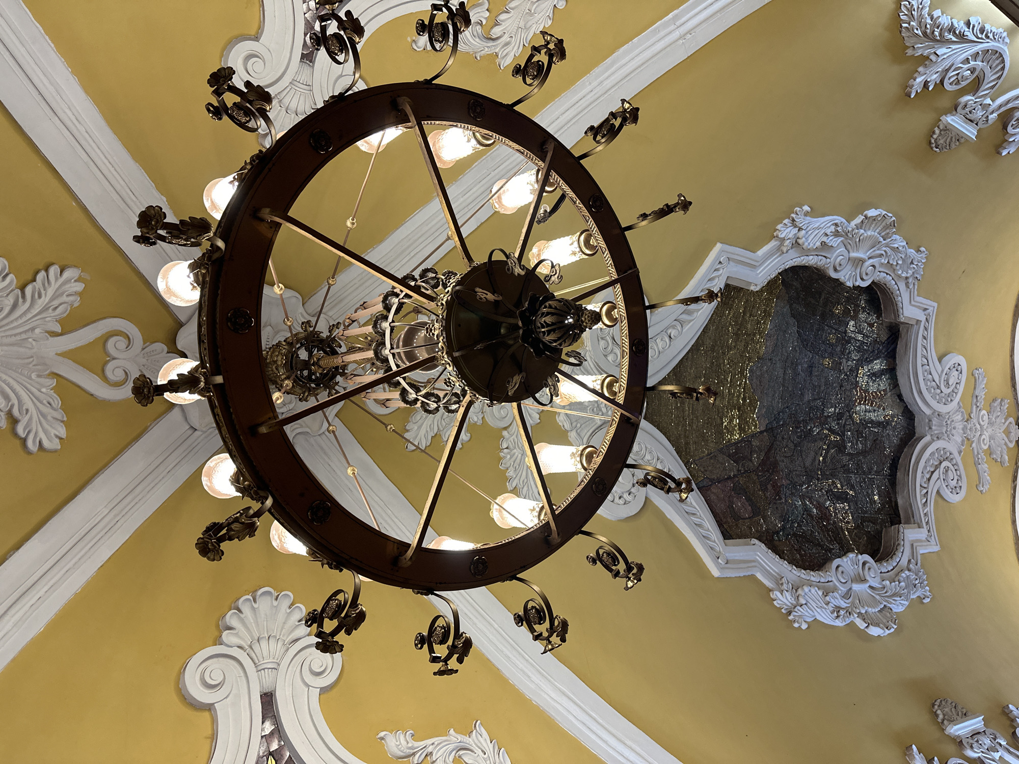 The image captures a view looking upwards towards a ceiling adorned with ornate decorative elements, predominantly in yellow and white, with a large, circular chandelier as the central focal point.
The ceiling features intricate white plaster moldings that frame sections painted in a muted yellow color. These decorations appear to be in a baroque or rococo style, with swirling patterns and floral motifs. In one section, the molding frames a mosaic panel of a dark scene of people, possibly within a building, or some other interior.
Hanging centrally is a substantial chandelier. It is structured as a dark brown wheel with numerous light bulbs mounted around its circumference. The lighting fixtures are suspended on curved metal arms that extend outwards from the wheel. The chandelier has a complex, multi-tiered center.
Based on the architectural style and decorative elements, this image likely depicts the interior of a Moscow Metro station. These stations are known for their lavish designs.