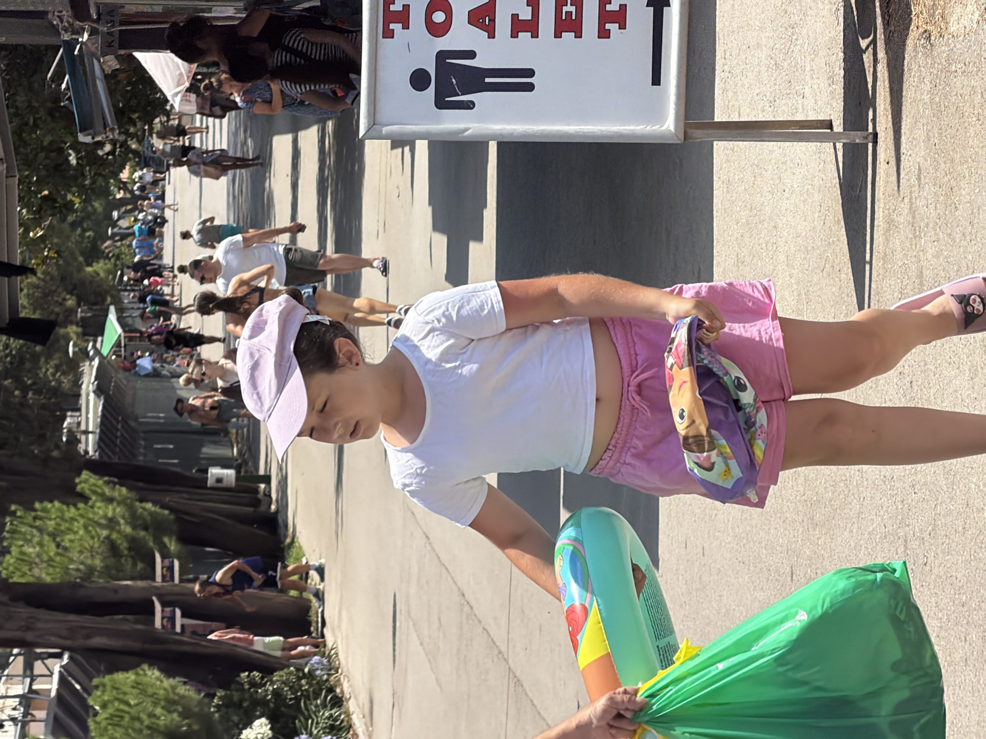The image depicts a scene in Budva, Montenegro, likely near a beach or promenade. In the foreground, a young girl wearing a white t-shirt and pink shorts is walking. She has a purple baseball cap on her head and is carrying a green plastic bag in her left hand and a colorful inflatable ring in her right hand. Next to her, a sign for "TOILET" is visible, with a male figure icon and an arrow pointing to the right. In the background, other people are walking along a path lined with trees and market stalls. The overall atmosphere suggests a sunny day in a tourist area.