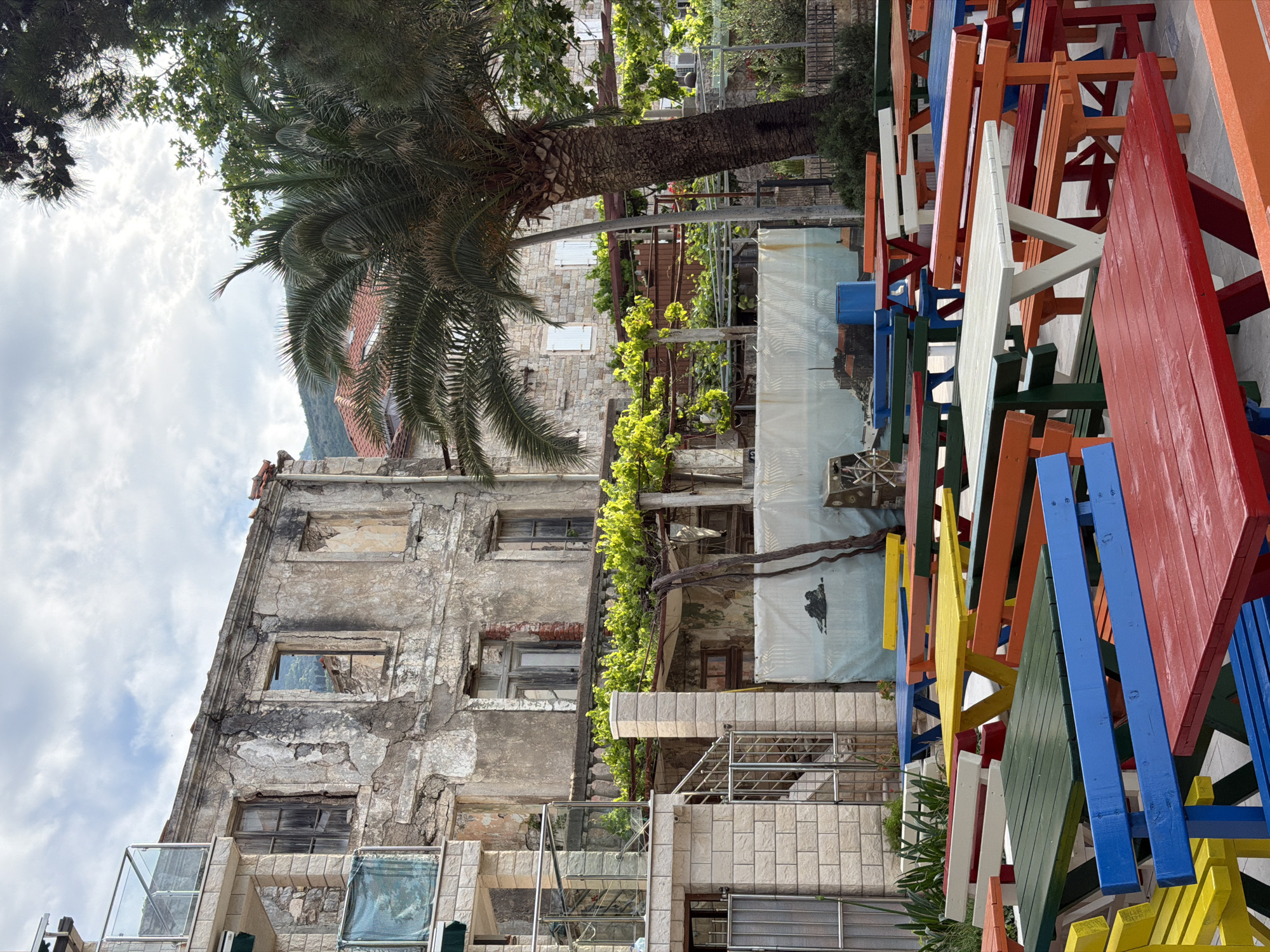 In Petrovac centar, Montenegro, an outdoor scene presents a weathered, multi-story building with visible signs of disrepair, including crumbling facade, broken windows, and missing sections. A large palm tree grows adjacent to the structure. Vegetation, including vines and bushes, is present. Stacked picnic tables, painted in bright colors such as red, blue, yellow, white, orange, and green, occupy the right side of the frame. The sky is overcast, with a cloudy background. The scene conveys a contrast between the decaying architecture and the vibrant, modern elements of the painted tables.