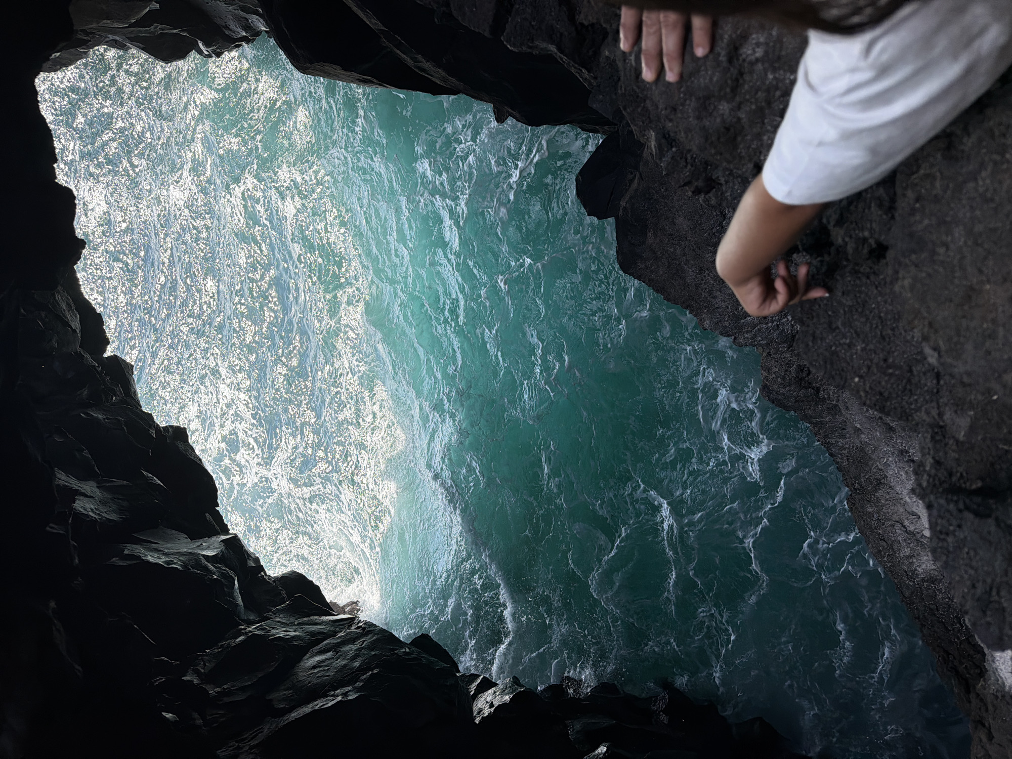 The image shows an ocean view through a cave opening. The turquoise water is visible with white foamy waves. Dark, rocky cliffs frame the opening on either side. A person's hand, arm, and shoulder are visible on the right side of the frame, positioned on the rock. The person is wearing a white sleeve. This is Los Hervideros in Yaiza, Lanzarote, Spain.