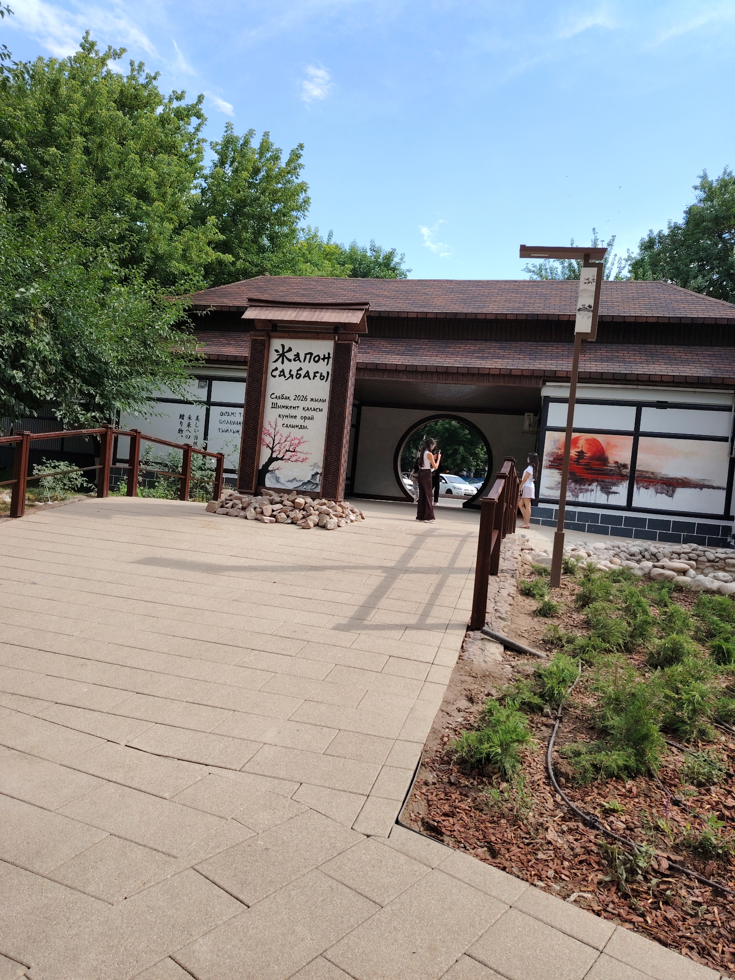 This is a medium-angle, eye-level, full outdoor shot of a building entrance with a circular opening in the facade. The building appears to be a Japanese-style restaurant or cultural center, with a dark brown tiled roof and dark wooden accents. In front of the building, there is a paved walkway made of light brown rectangular tiles. To the left of the walkway is a wooden fence and some green trees. To the right, there are some small evergreen bushes planted in bark mulch, and a light pole with a small banner.
A large sign with an illustration of a blooming cherry tree and Japanese calligraphy is positioned to the right of the entrance. There are two women near the entrance. One is standing inside the circular opening, facing away from the camera and holding a phone up, possibly taking a picture. The other woman is standing just inside the building, to the right of the circular opening, facing the camera and looking down. On the wall of the building to the right of the entrance, there is a mural of a red sun setting behind a Japanese pagoda.
The sky is blue with some scattered white clouds. The overall impression is of a serene and aesthetically pleasing environment, likely a public space or establishment. The location is identified as Unknown, Unknown.