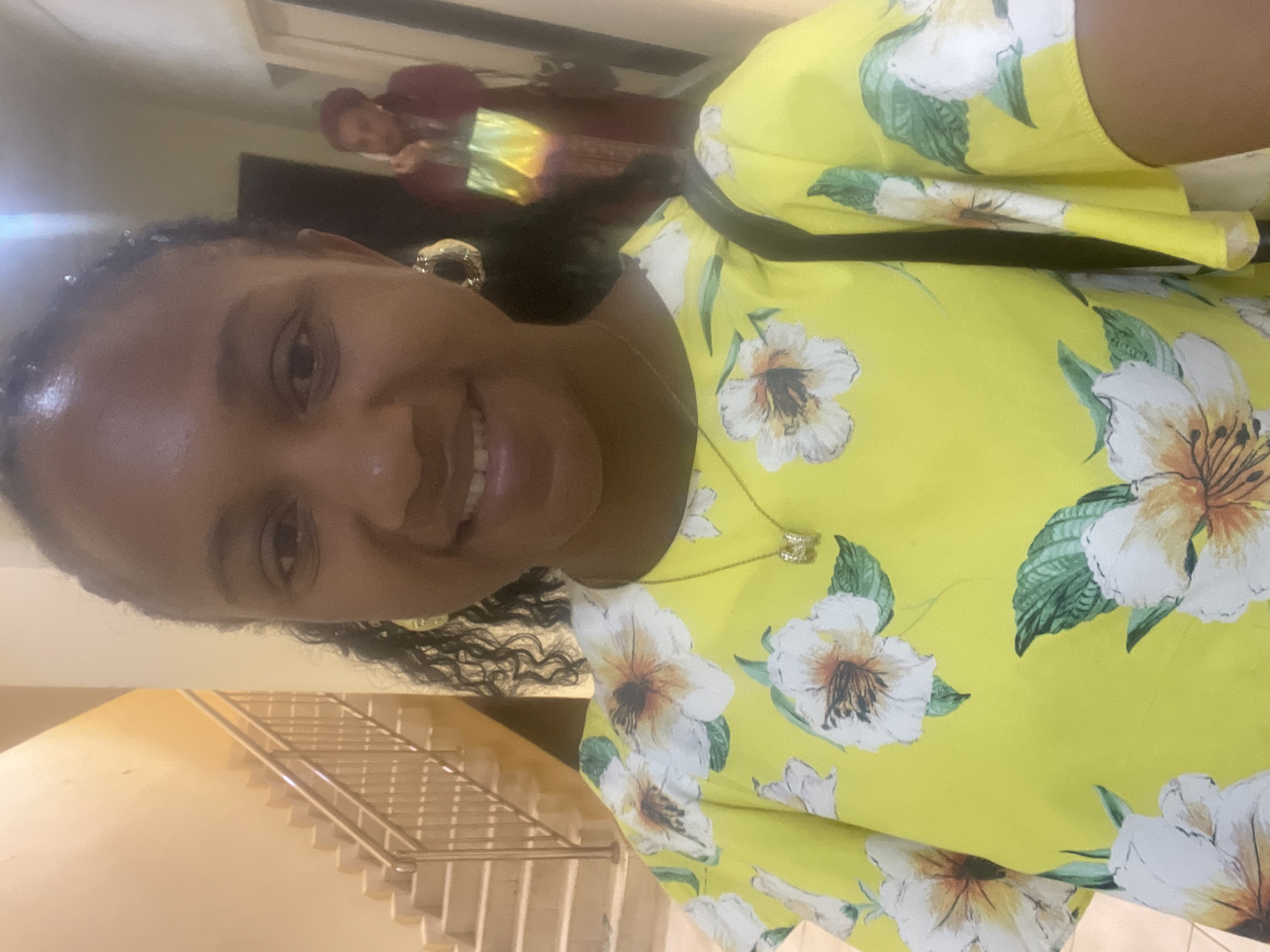 A woman smiles into the camera, wearing a yellow floral top, gold earrings, and a gold necklace. She appears to be standing indoors near a staircase with metal railings. In the background, a person is partially visible. The location is potentially the city of Jos, Nigeria.