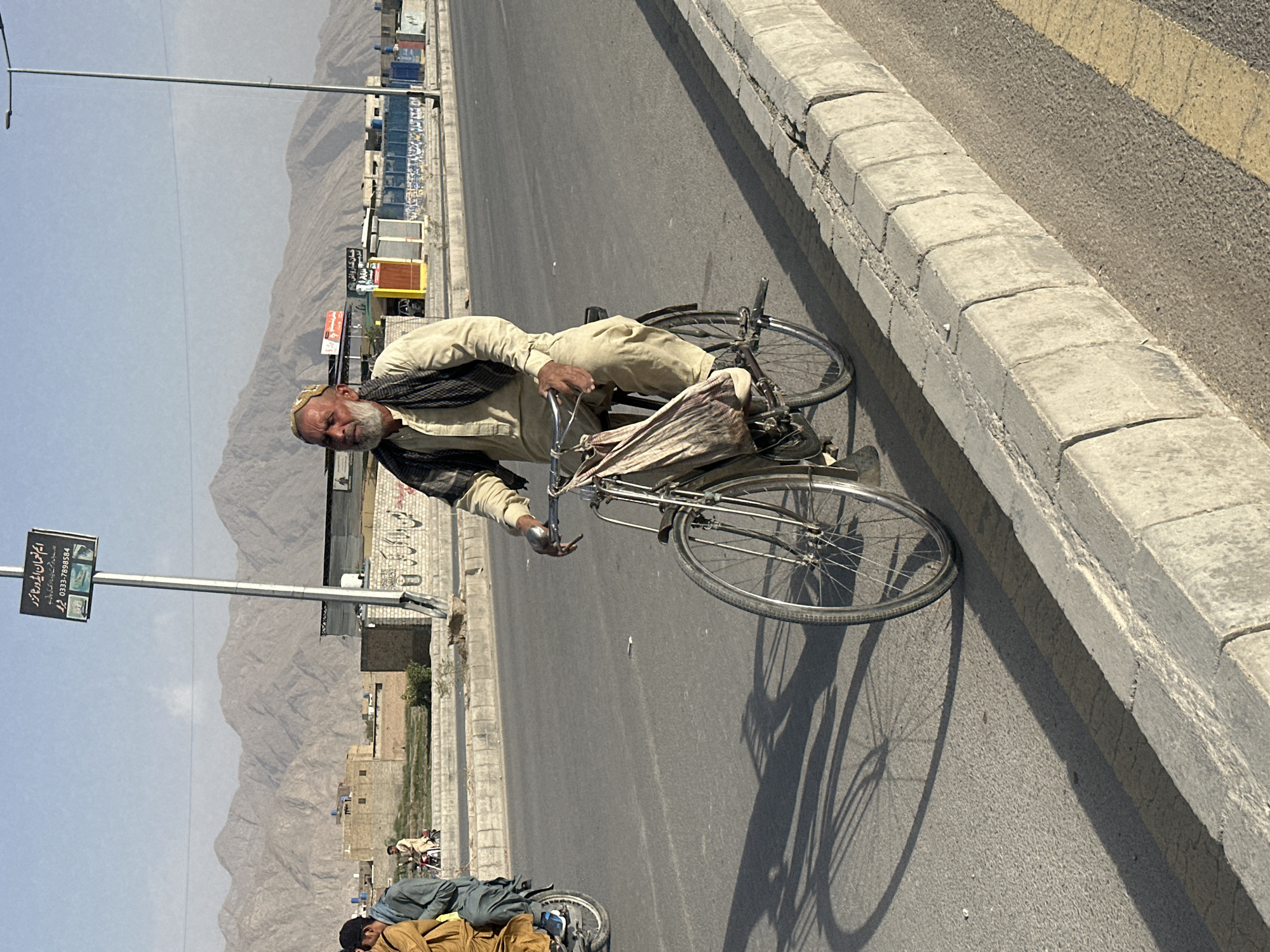 An elderly Pashtun man with a white beard and a patterned skullcap is riding a bicycle on a paved road in Quetta City Tehsil, Pakistan. He is wearing a light-colored kameez and shalwar with a dark vest. A cloth bag is hanging from the bicycle's handlebars. The man is riding on the right side of the road, and there is a grey concrete curb to his right. In the background, there are buildings and mountains. The sky is clear and blue. There are shadows of the cyclist and bicycle on the road.