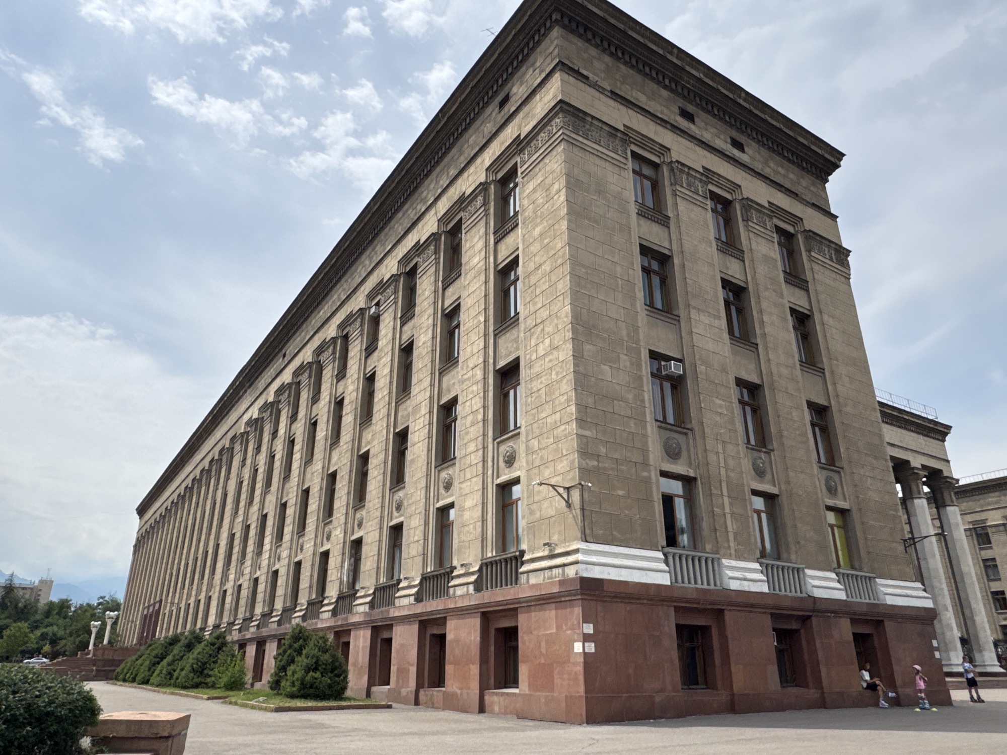 The image depicts the **Kazakhstan National Academy of Sciences** building, a large, imposing structure in Almaty, Kazakhstan. The building is made of light-colored stone blocks and features numerous regularly spaced windows. It has a distinctive architectural style, with ornate detailing along the upper facade and a red granite base. The perspective is from a low angle, looking up at the building, emphasizing its scale. The sky is partly cloudy, with a soft, diffused light.
In the foreground, on the paved area in front of the building, are a few individuals. To the right, two children are present: one is wearing a pink hat and appears to be on roller skates or a similar wheeled device, while another, dressed in white, is also on roller skates or a similar device. Further to the right, a third person, wearing dark shorts and a white shirt, is standing. In the middle ground, to the left of the building, a line of green bushes and trees separates the building from the street. A few white pillars and steps are visible leading up to an entrance or a different section of the complex. A white car is parked to the left of the bushes. The overall scene suggests a public space in front of a significant institutional building.
