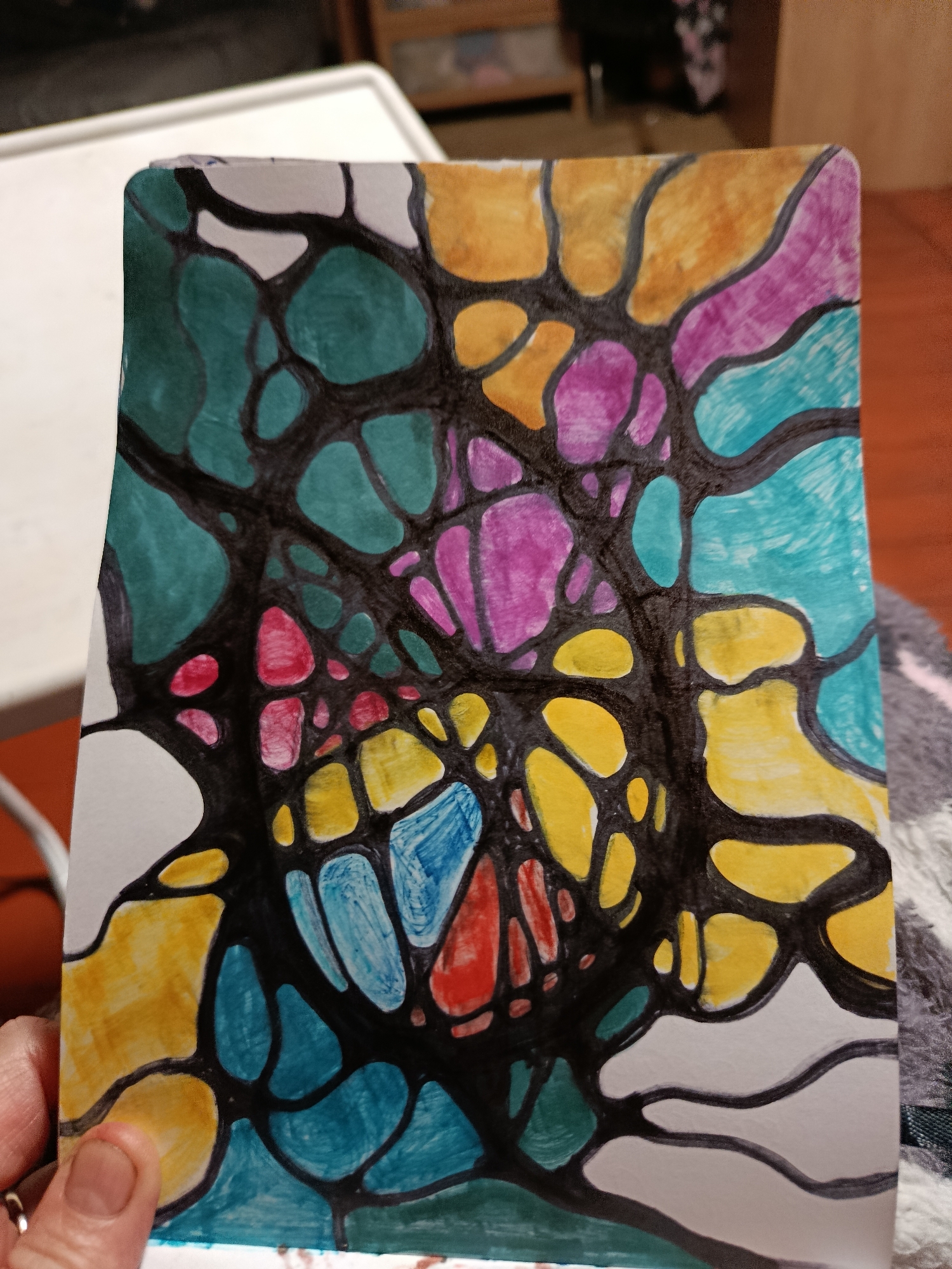 The image is a close-up, overhead shot of a colorful drawing on a piece of white paper. The drawing features a network of thick black lines that create a series of interconnected, organic shapes. These shapes are filled with various vibrant colors including teal, yellow, pink, purple, red, and blue. The paper appears to be a postcard or a small card, with slightly rounded corners. The drawing is held by a person's hand, with a thumb and finger visible in the lower left corner. A ring is visible on the index finger. The background of the image is blurred, showing a white surface, possibly a table, and a wooden surface, likely furniture, in the distance. The lighting appears to be indoor lighting. The scene is not in a city, but rather in an indoor setting.