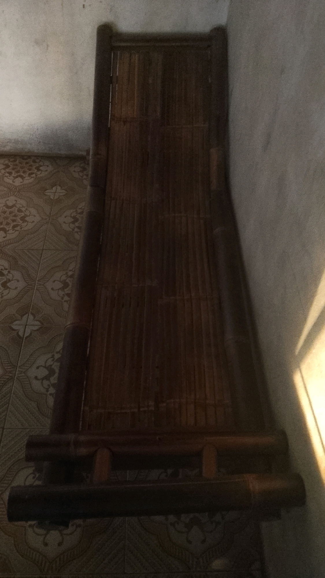 In the city of Mulyo Haji, Indonesia, a dark brown, woven bamboo cot rests on a beige tiled floor. The cot is positioned at an angle in a corner, with its head against a light-colored, textured wall. The floor tiles feature a repeating pattern of light brown floral motifs. A shadow cast by an unseen object or light source falls across the right side of the wall and the cot, creating a diagonal line of light and dark. The scene is dimly lit, with the majority of the cot and the surrounding area in shadow.